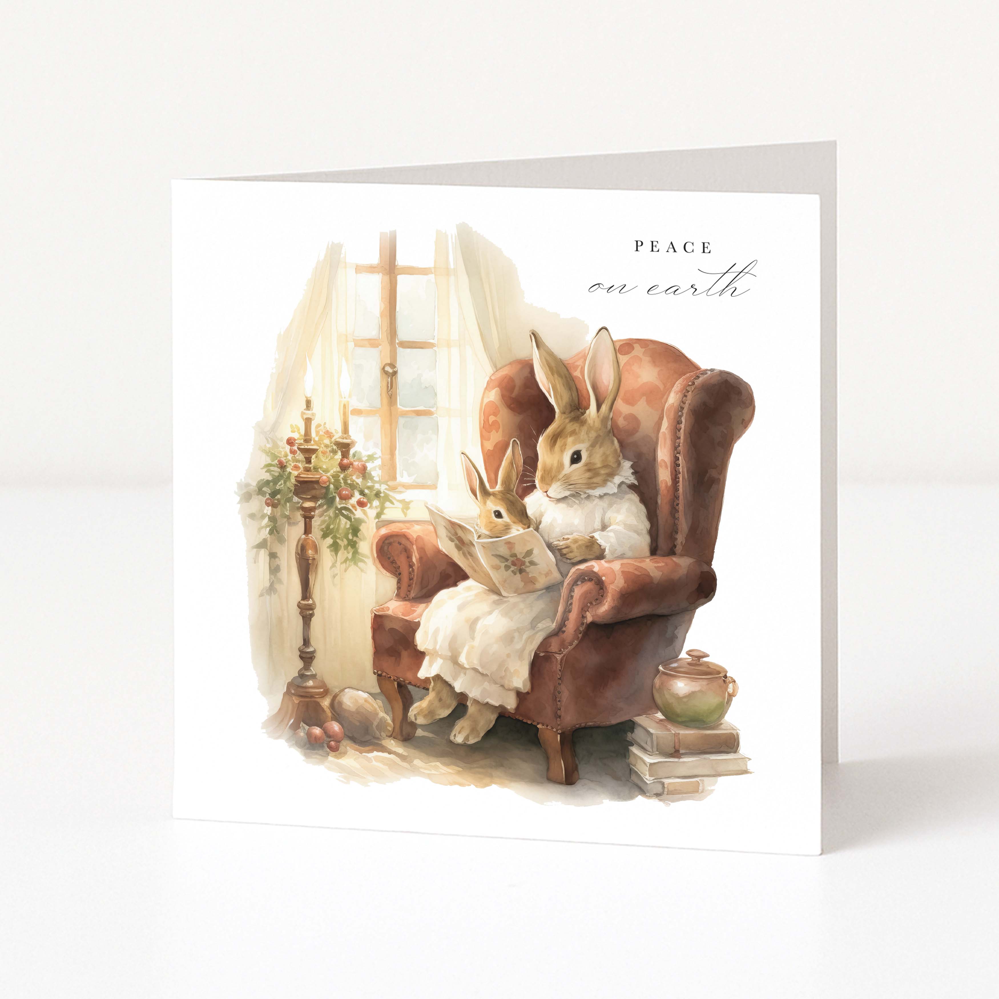 A square Christmas greeting card shown standing upright on a white surface. The front features a watercolour-style illustration of two rabbits sitting in a brown armchair beside a window, with the words 'PEACE on earth' printed near the top right.