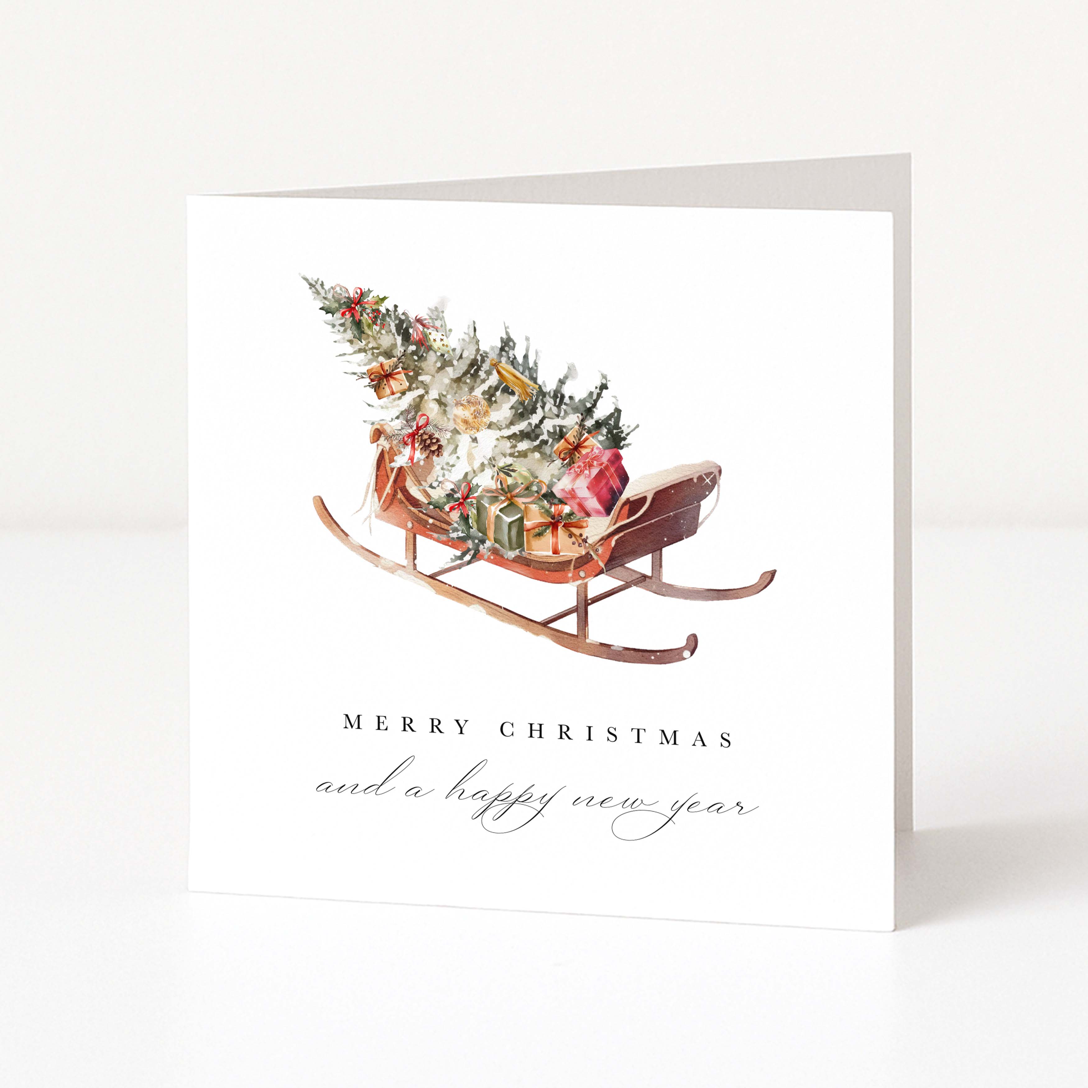 A white Christmas greeting card standing upright, featuring a watercolor illustration of a sleigh carrying a decorated Christmas tree and wrapped gifts, with the text 'MERRY CHRISTMAS and a happy new year' below.