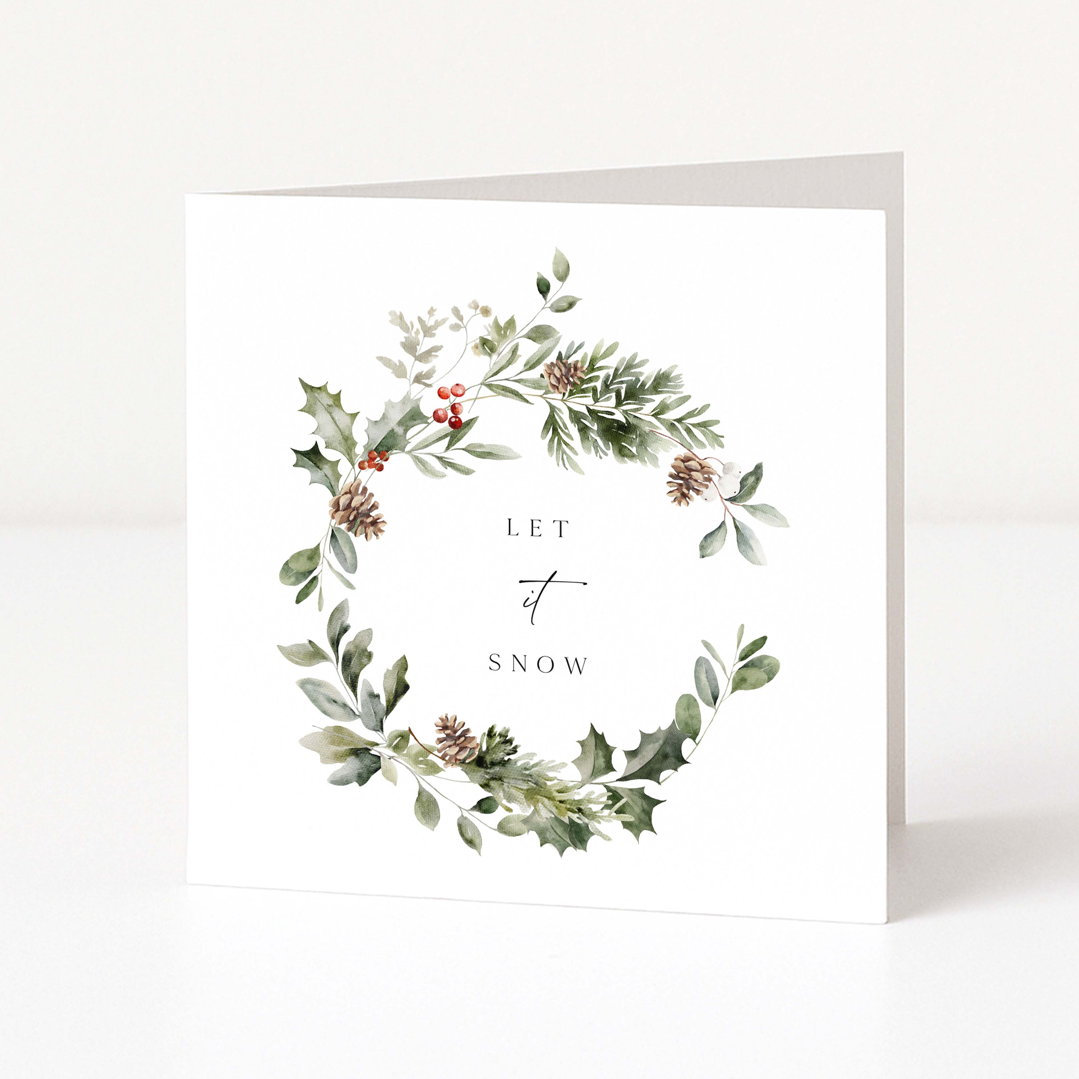 A square white greeting card with a watercolour wreath of green foliage, pinecones, and small red berries around the words 'LET IT SNOW' in black serif and script text, shown standing against a clean white background.