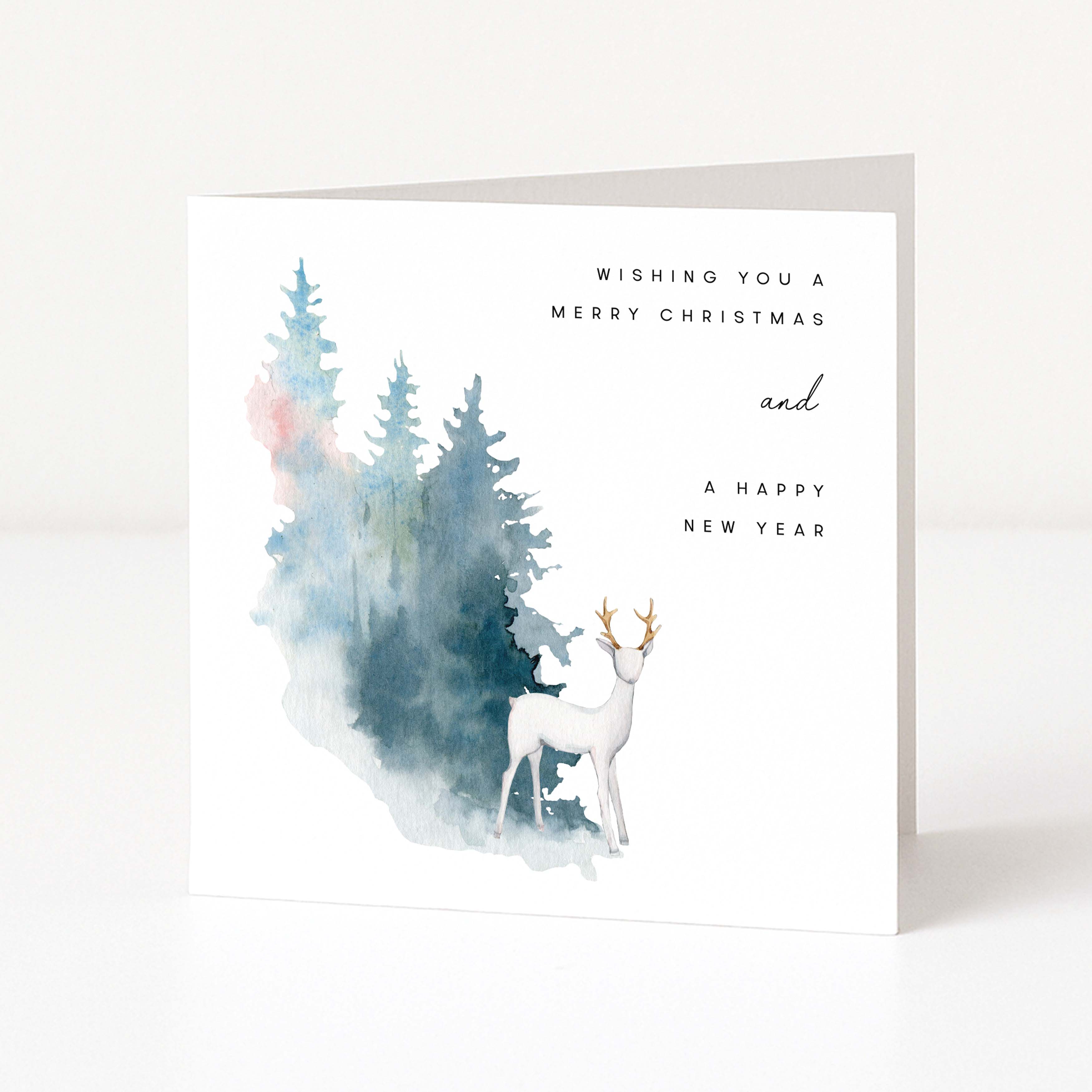 A square white greeting card standing upright on a light grey surface. The front features a watercolour-style forest with blue and green trees, a white deer with gold antlers, and the text 'Wishing you a Merry Christmas and a Happy New Year'.