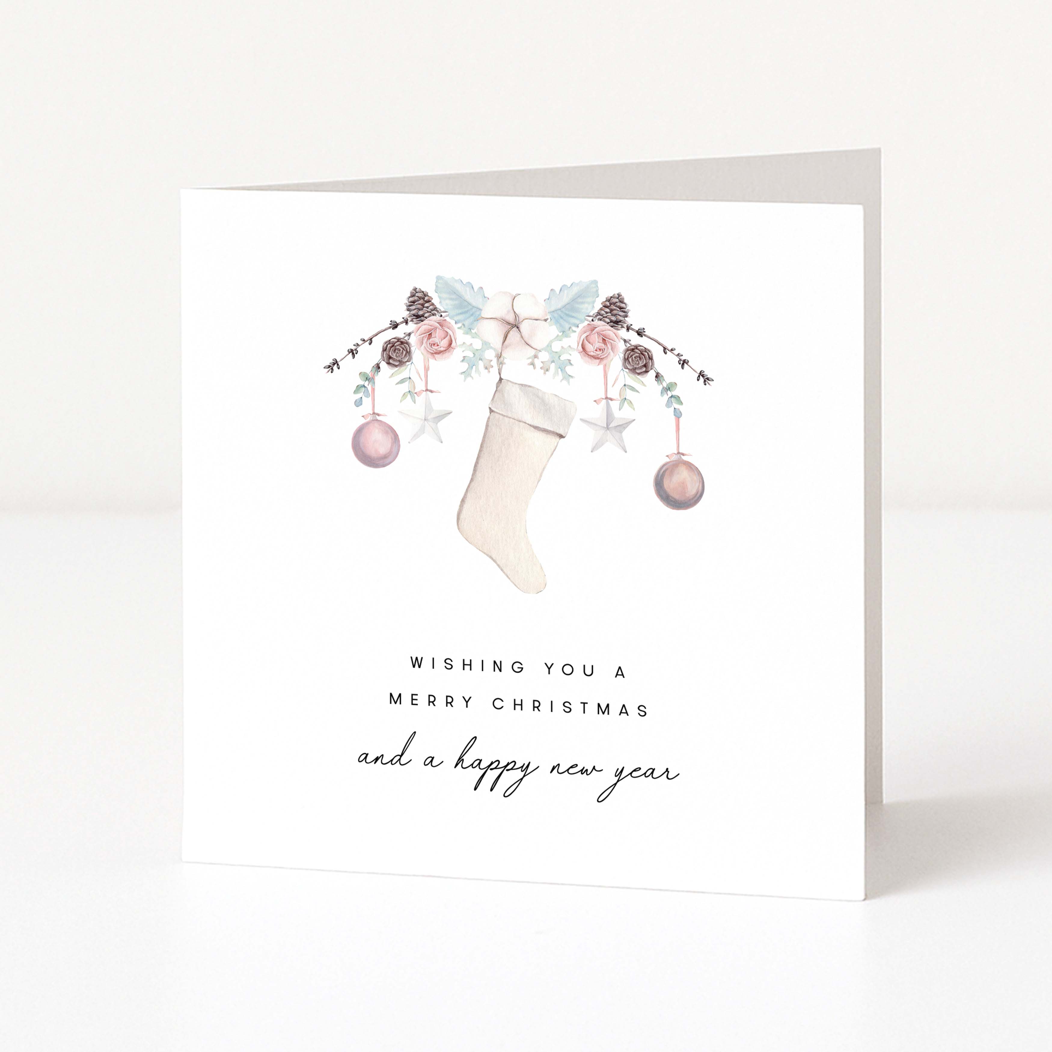 A white greeting card standing upright on a light surface, featuring a watercolour Christmas stocking decorated with pastel flowers, pinecones, and ornaments, with the text 'Wishing you a Merry Christmas and a happy new year'.