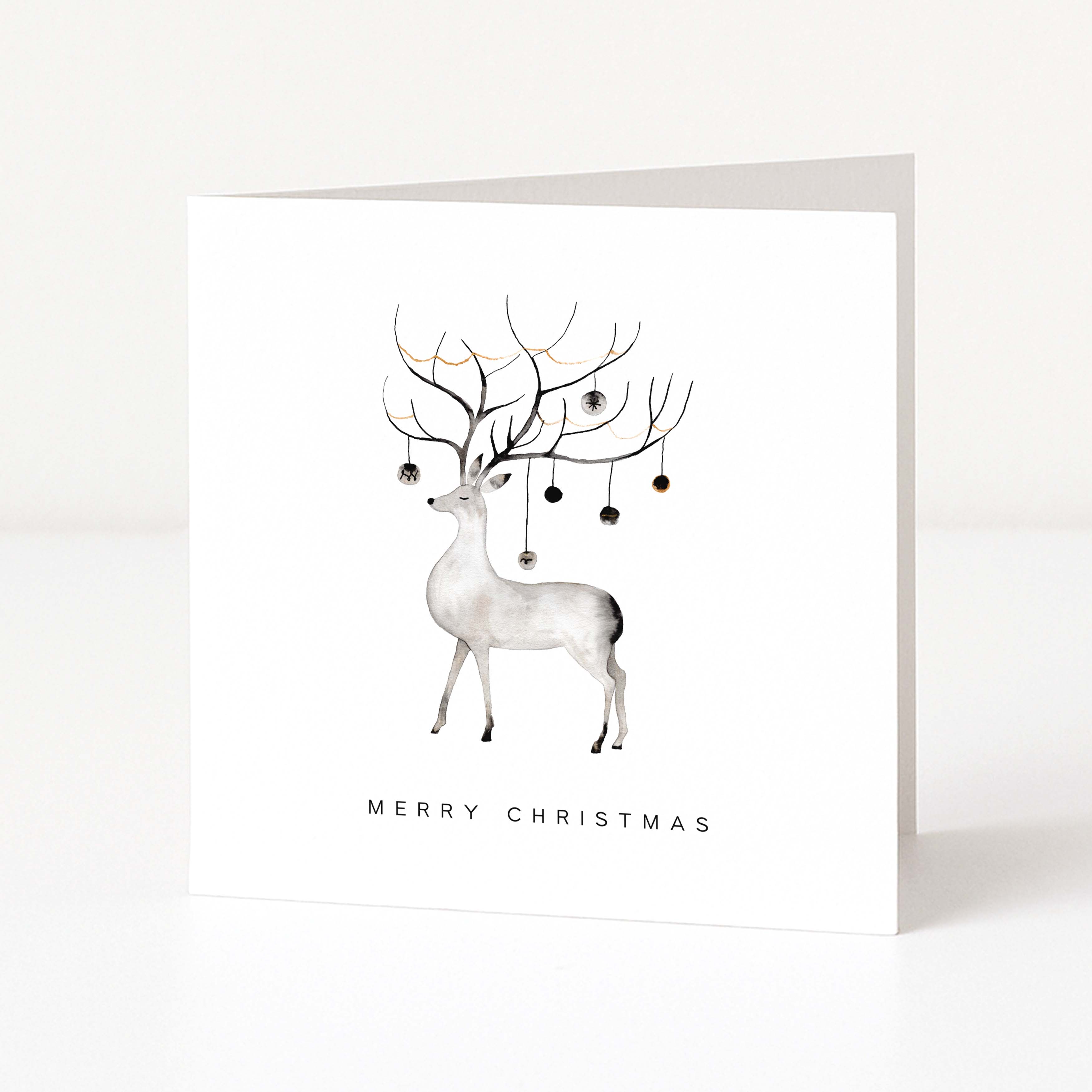 A white square Christmas greeting card standing upright on a white surface, featuring a minimalist illustration of a white deer with antlers decorated with small dark ornaments and the text 'MERRY CHRISTMAS' below.