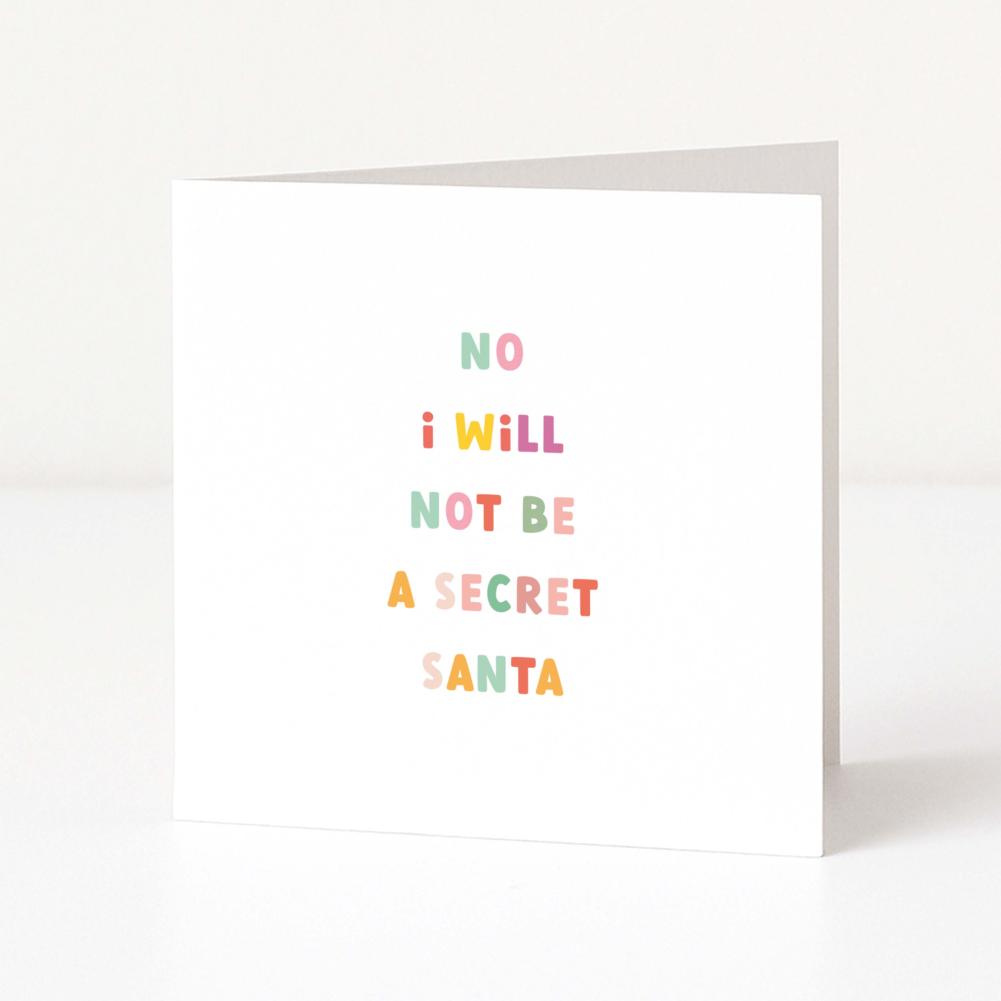 A white anti-Christmas card standing upright on a white surface, with the phrase 'NO I WILL NOT BE A SECRET SANTA' printed in small pastel letters.