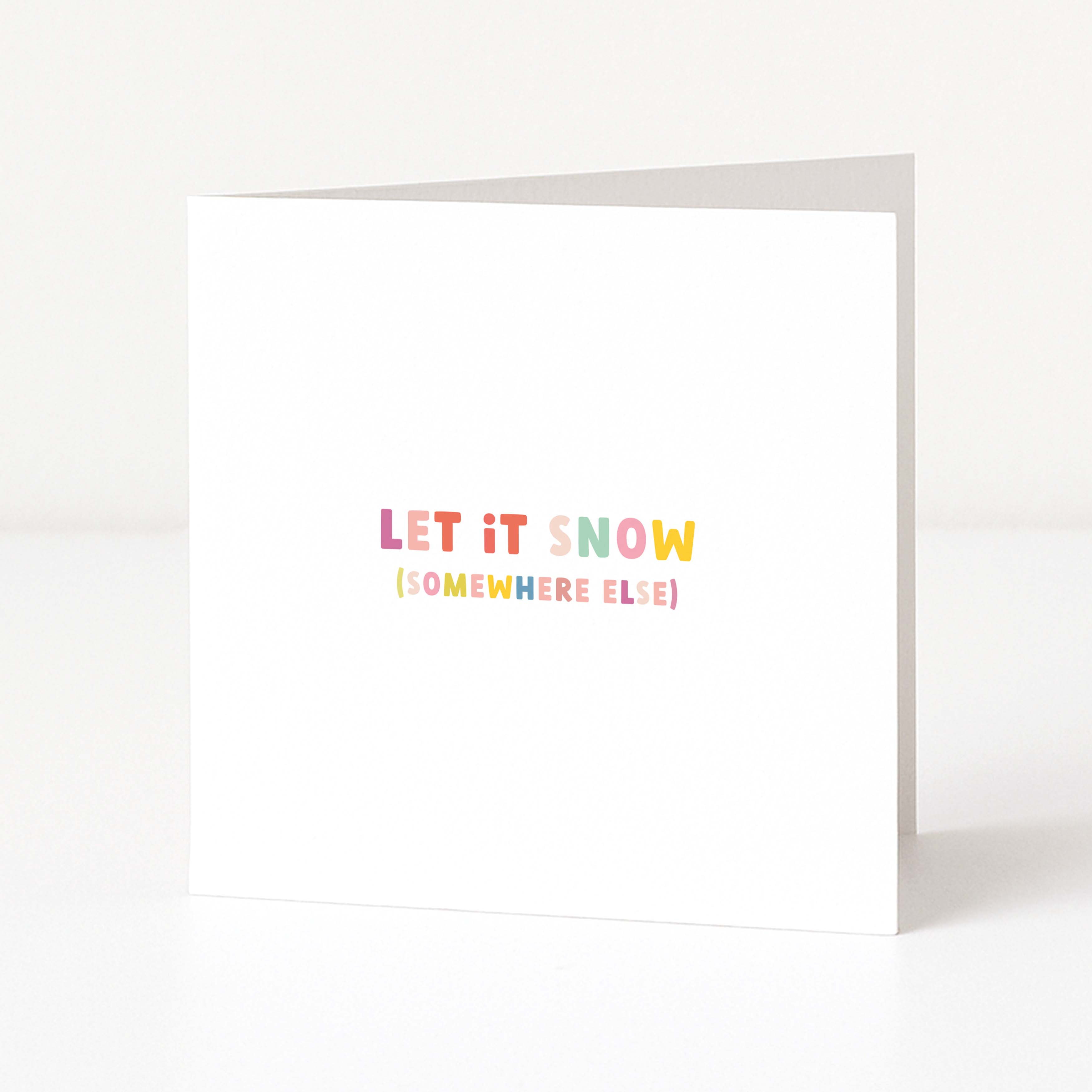 A white anti-Christmas card standing upright on a white surface, with the phrase 'LET IT SNOW (SOMEWHERE ELSE)' printed in small pastel rainbow letters near the center.