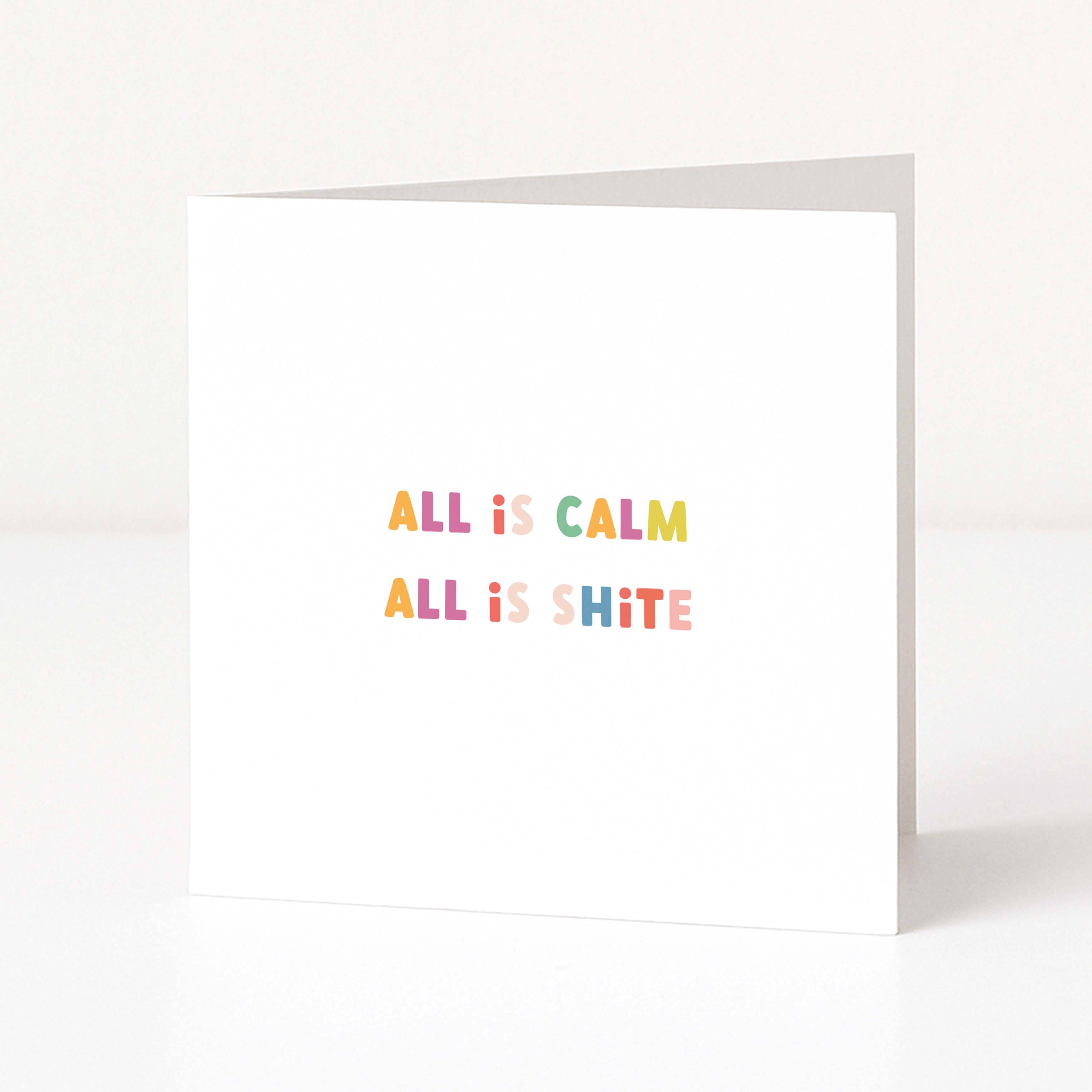 A white anti-Christmas card standing upright on a white surface, with the phrase 'ALL IS CALM ALL IS SHITE' printed in small pastel letters across the centre.