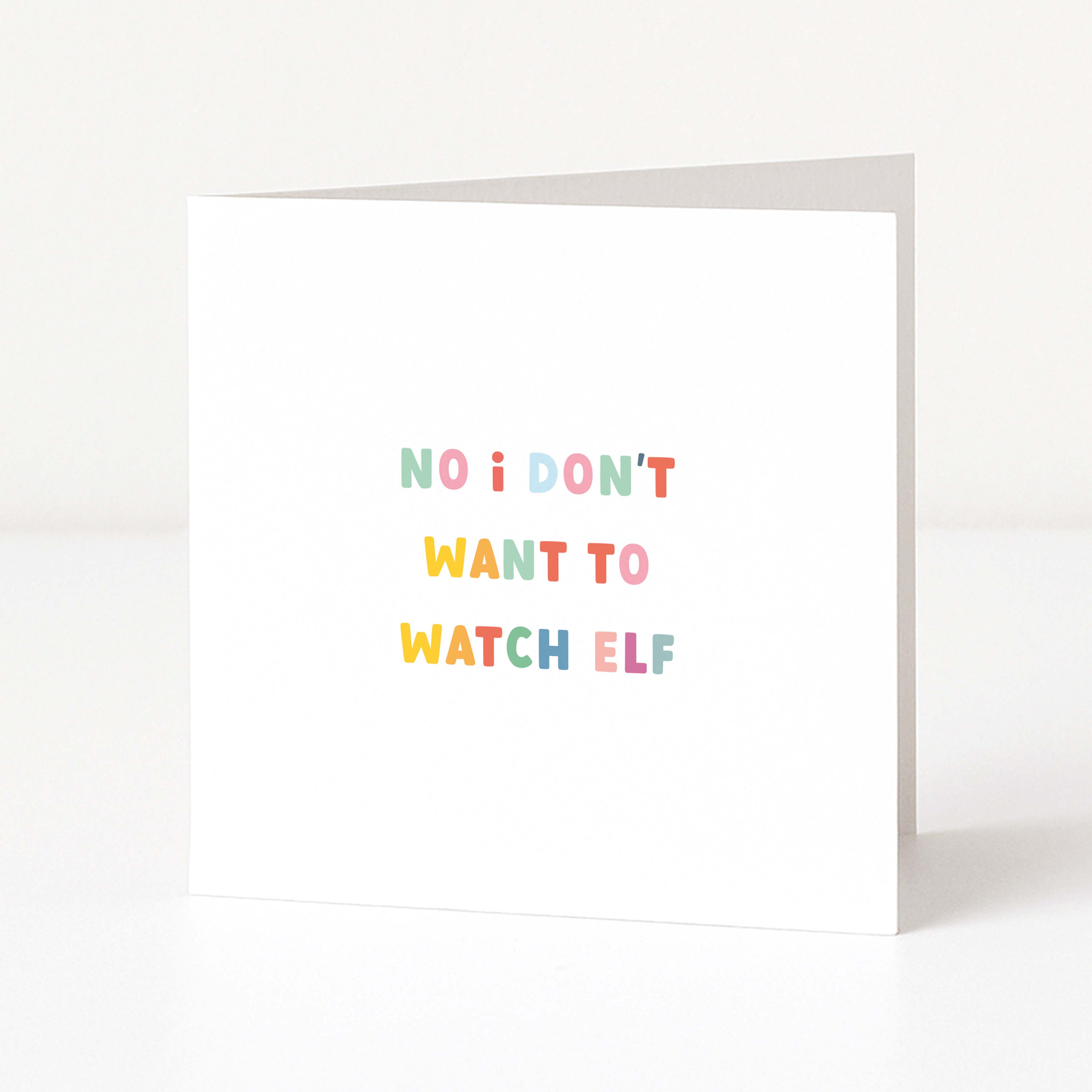 A white anti-Christmas card standing upright on a white surface, with the text 'NO i DON'T WANT TO WATCH ELF' printed in small pastel rainbow letters.