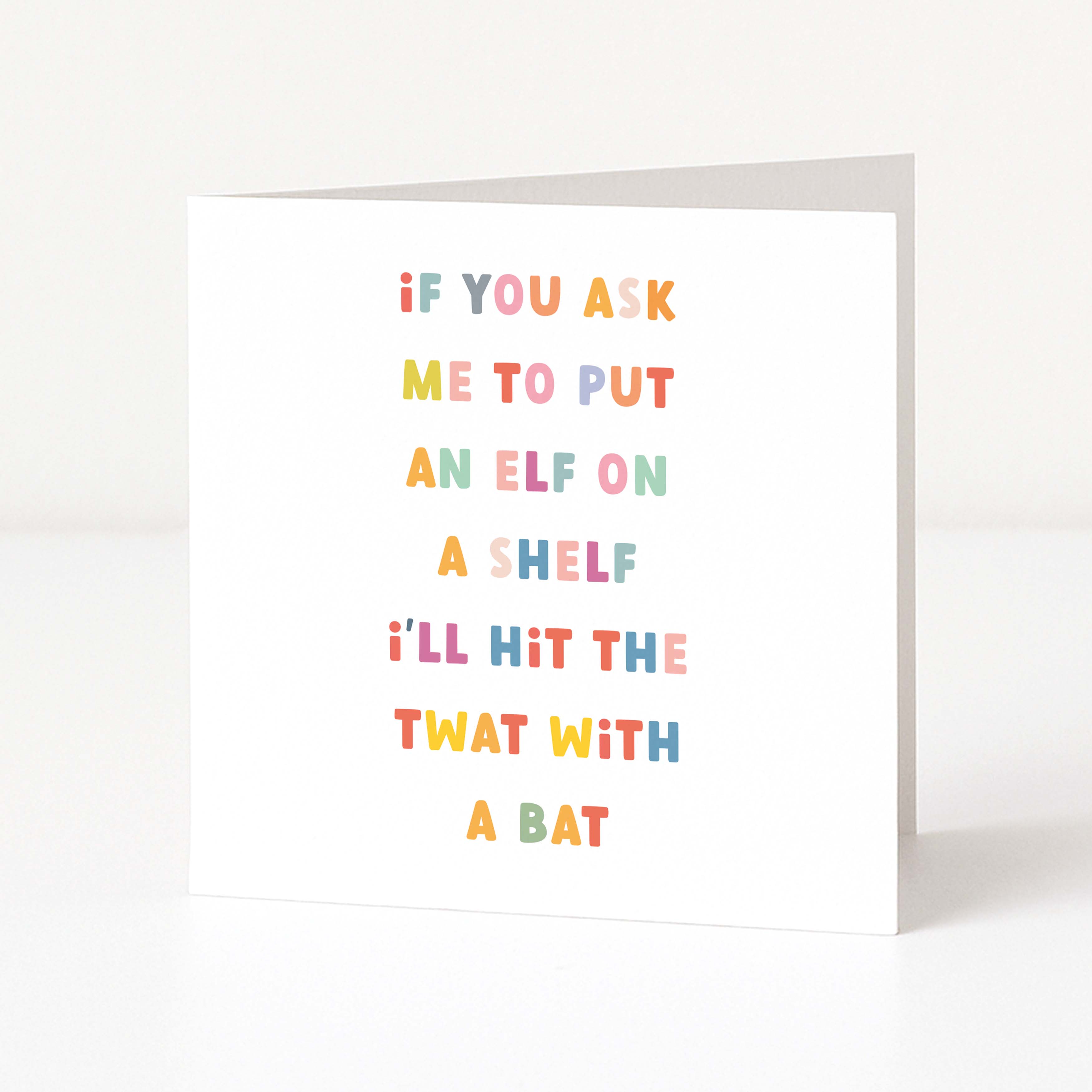 A white anti-Christmas card standing upright on a white surface, printed with the punny phrase 'IF YOU ASK ME TO PUT AN ELF ON A SHELF I'LL HIT THE TWAT WITH A BAT' in colorful pastel letters.