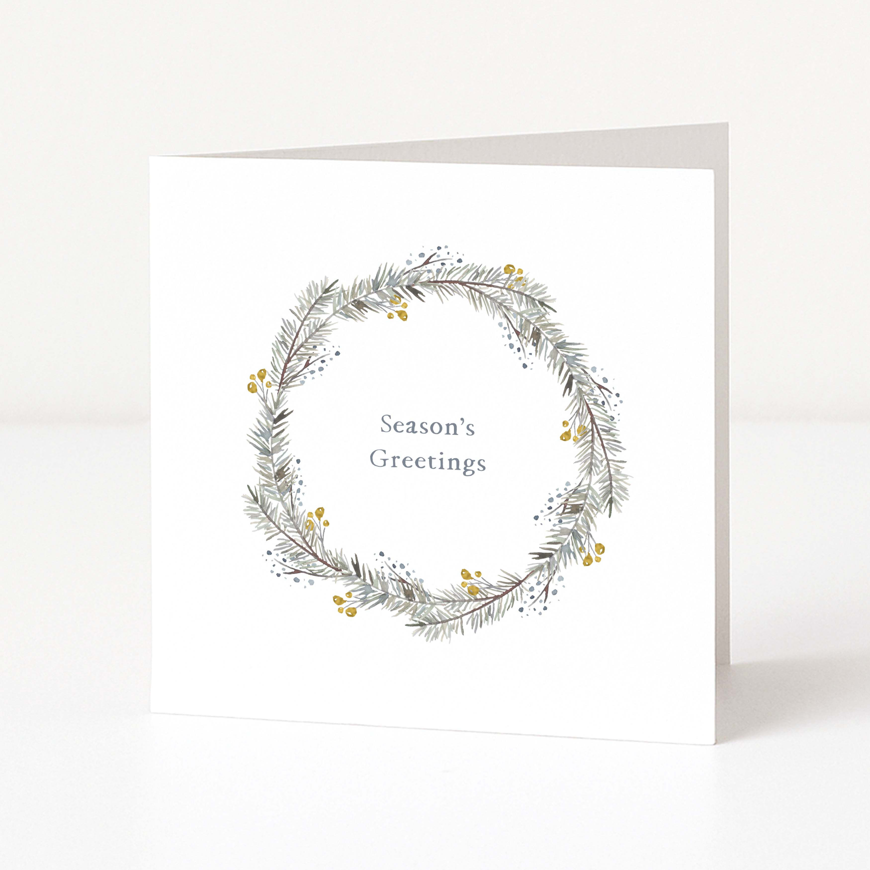 A white square Christmas greeting card standing upright on a light neutral surface. The front features a delicate watercolour wreath in muted green, grey, and gold tones with the words 'Season's Greetings' centred inside.
