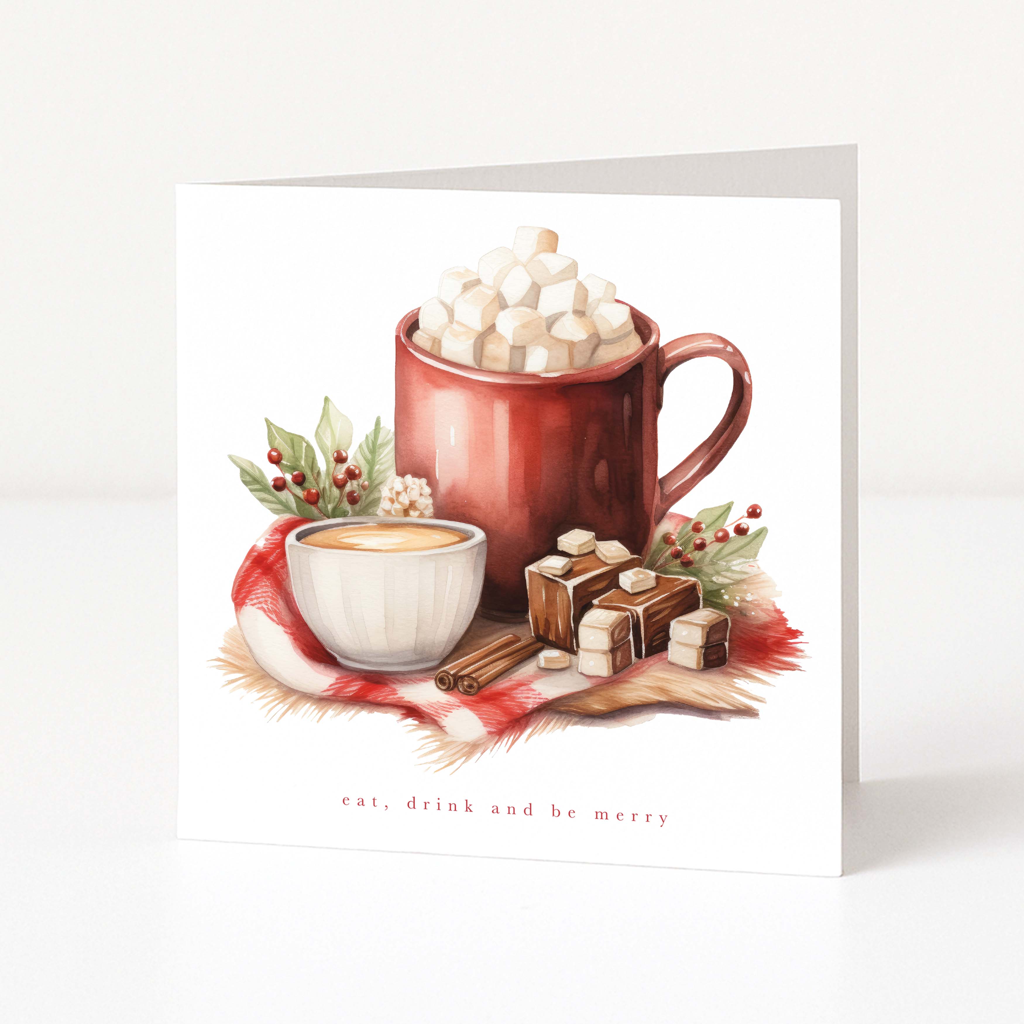 A square Christmas greeting card featuring a watercolour illustration of a red mug filled with marshmallows and hot chocolate, alongside a cup of tea, cinnamon sticks, and chocolate squares on a red plaid cloth. The card reads 'eat, drink and be merry' at the bottom, shown standing upright against a clean white background.