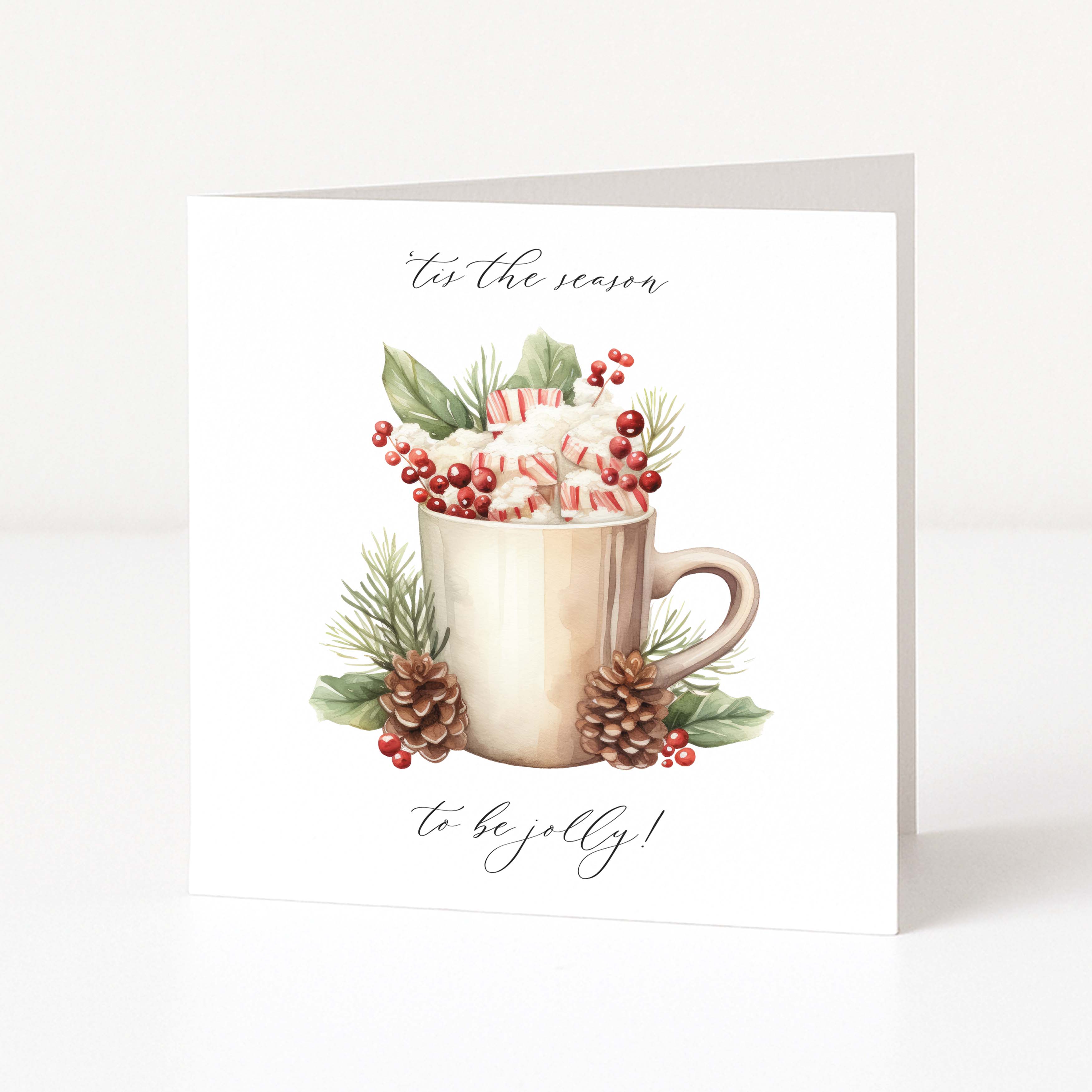 A square white Christmas greeting card standing upright on a light grey surface, featuring a watercolour-style illustration of a beige mug filled with whipped cream and candy canes, surrounded by green leaves, red berries, pinecones, and the phrases 'for the season' and 'to be jolly!'.