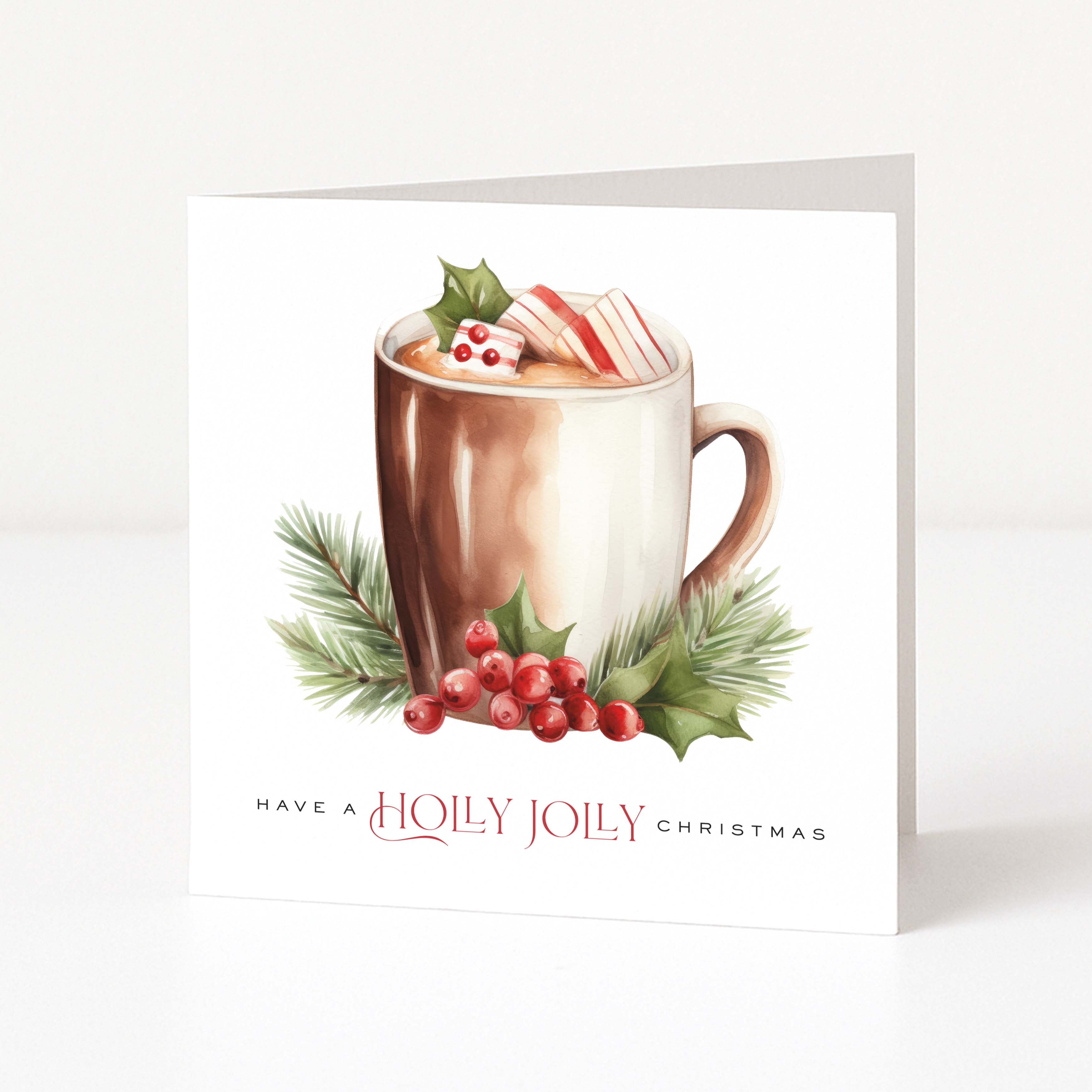 A square white Christmas greeting card standing upright on a light grey surface. The front features a watercolour-style illustration of a hot mug with cocoa, green holly leaves, red berries, and a striped candy cane, with the text 'HAVE A HOLLY JOILY CHRISTMAS' below.