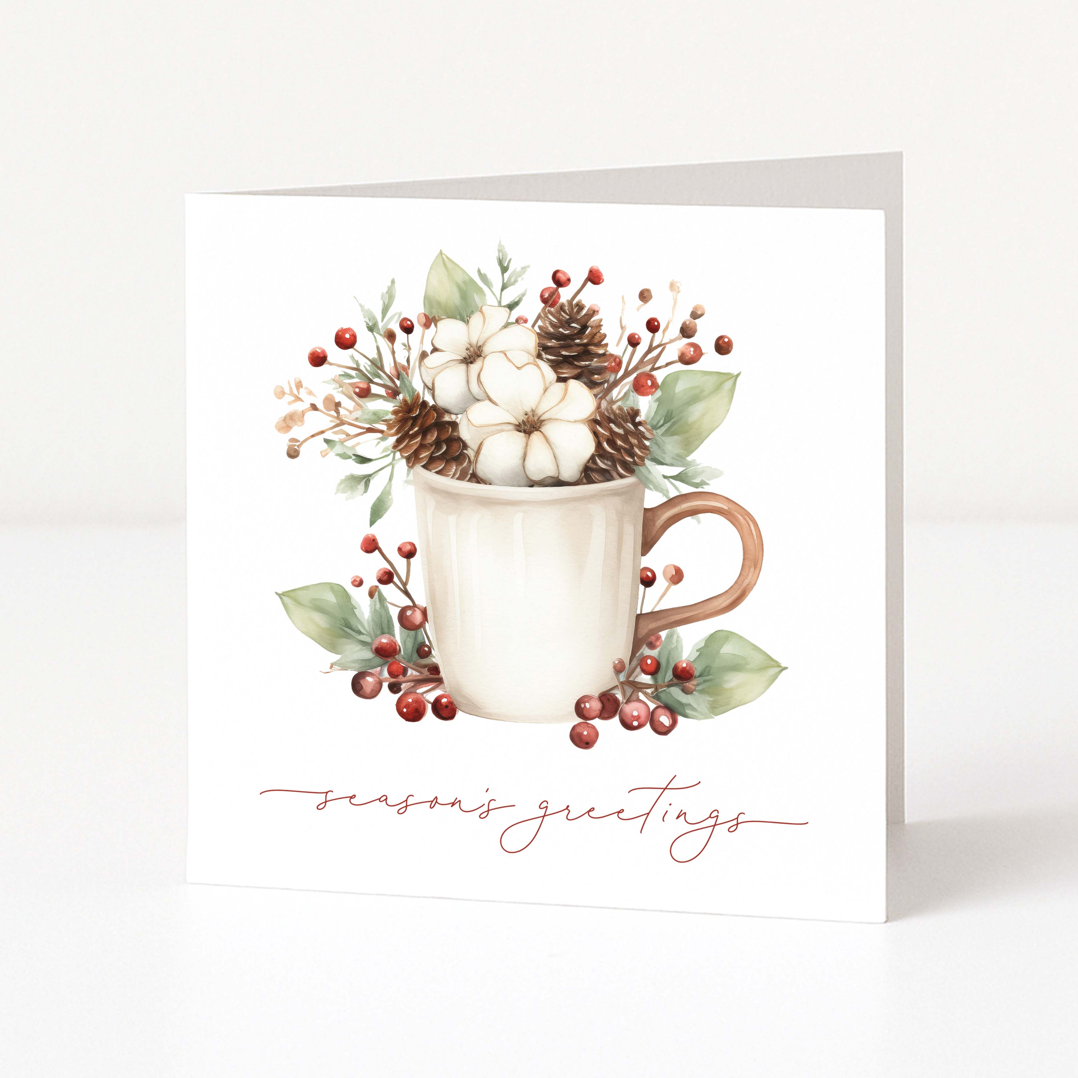 A white Christmas greeting card standing upright on a light surface, featuring a watercolour-style illustration of a white mug filled with cotton bolls, pinecones, green leaves, and red berries, with the words 'season's greetings' in a rose-gold script at the bottom.