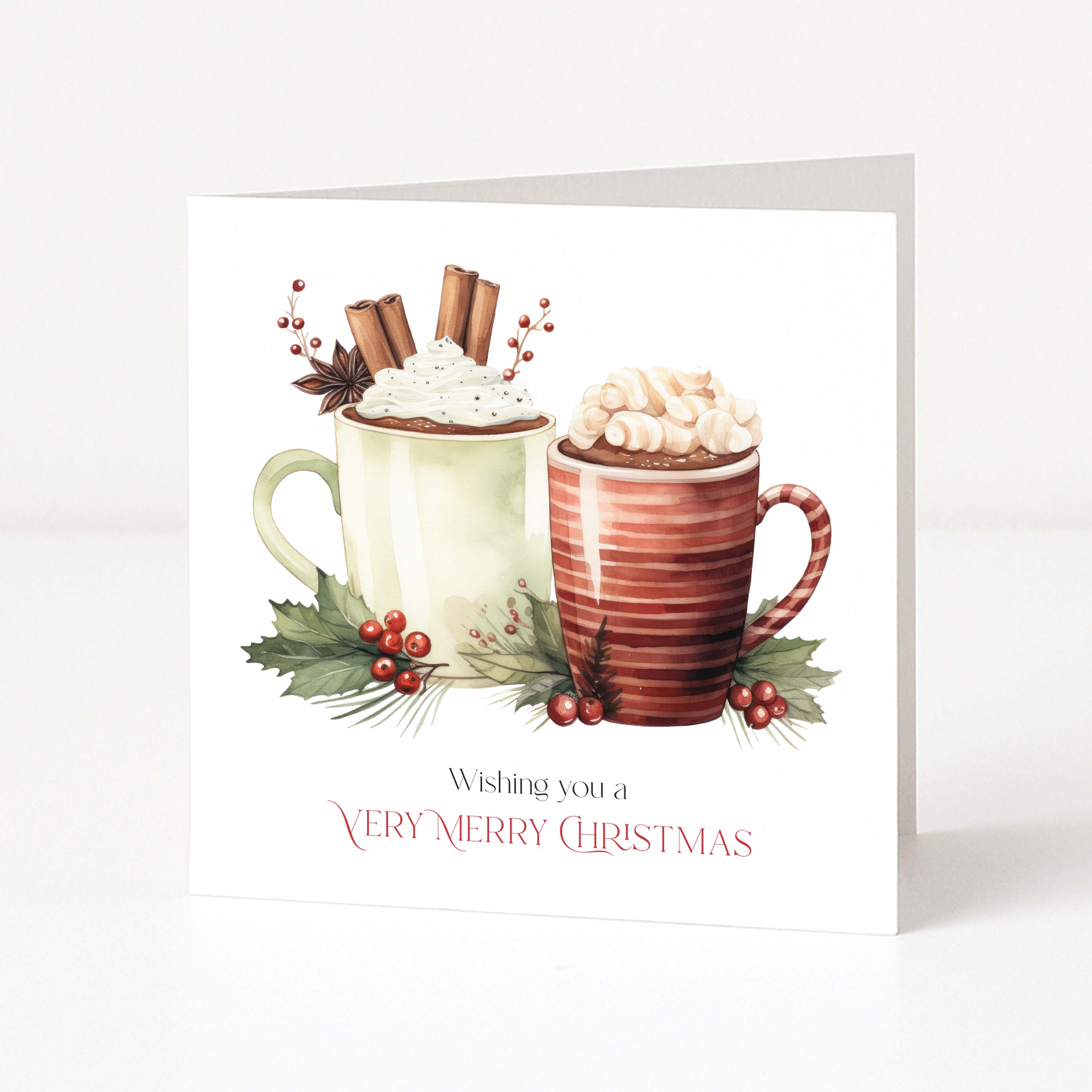 A square Christmas greeting card standing upright on a white surface. The card features an illustration of two hot cocoa mugs topped with whipped cream, with cinnamon sticks, star anise, holly leaves, and red berries.