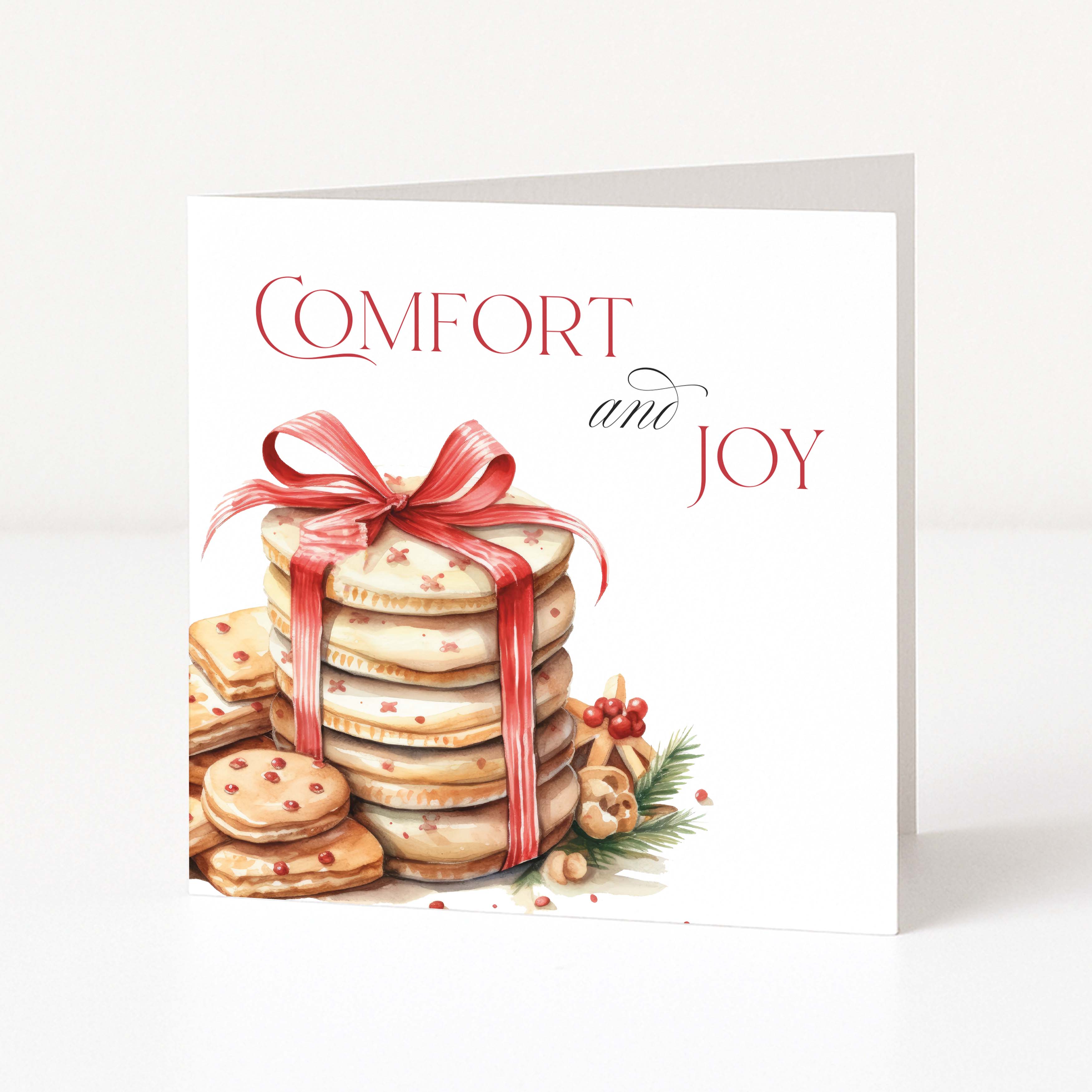 A white Christmas greeting card standing upright, printed with the words 'COMFORT and JOY' above an illustration of stacked holiday cookies tied with a red ribbon, on a clean white background.