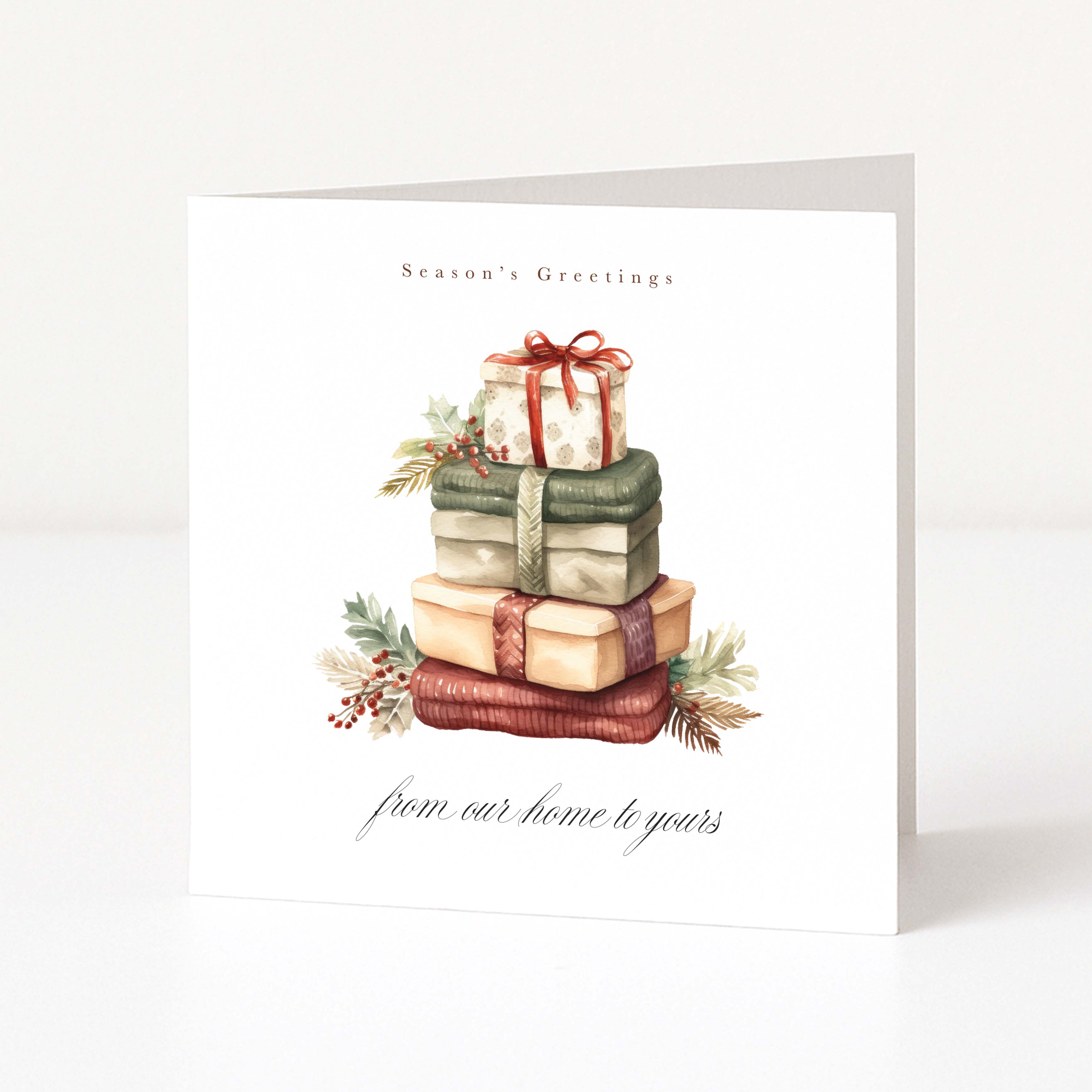 A white Christmas greeting card standing upright on a light surface, printed with the words 'Season's Greetings' and 'from our home to yours' and featuring an illustrated stack of wrapped Christmas presents with greenery accents.
