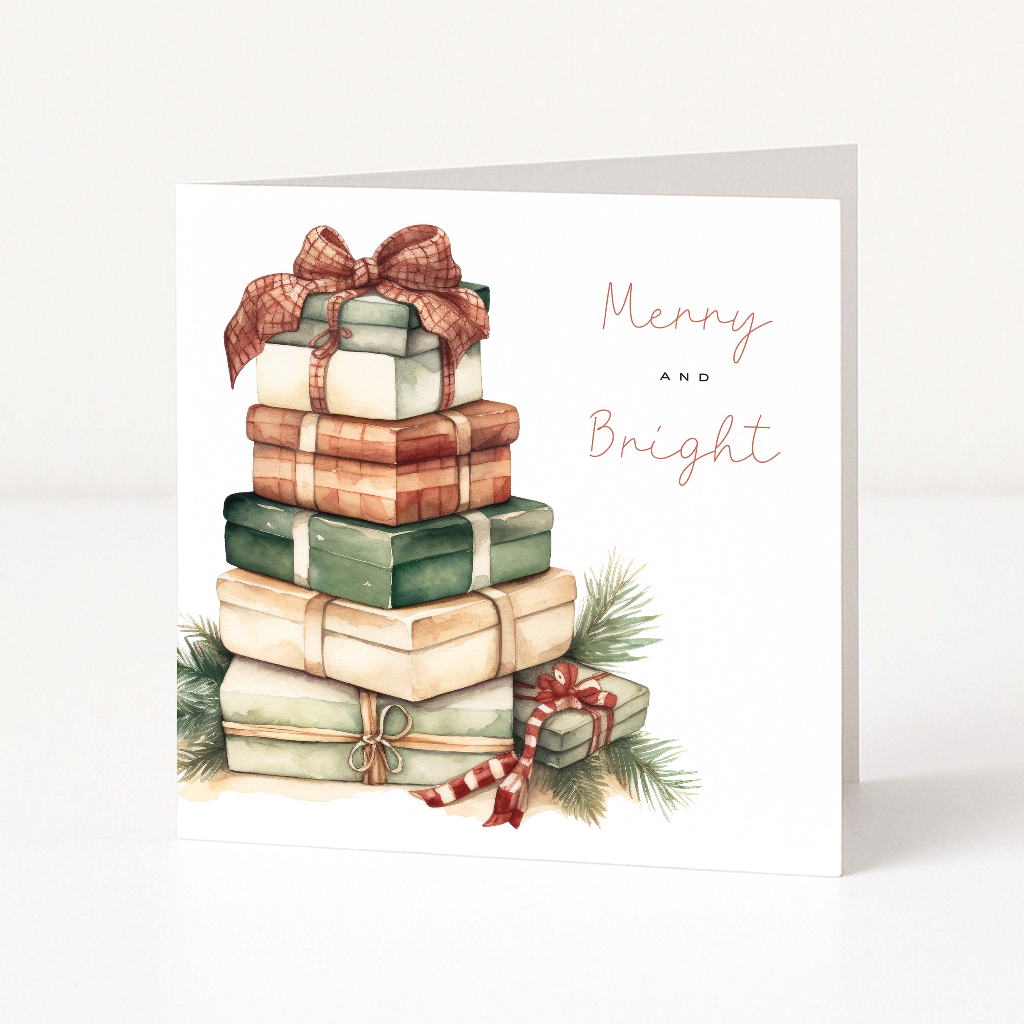 A white Christmas greeting card featuring a watercolour illustration of stacked gift boxes tied with ribbons, green pine branches, and the words 'Merry and Bright' in peach script, shown standing against a light neutral background.