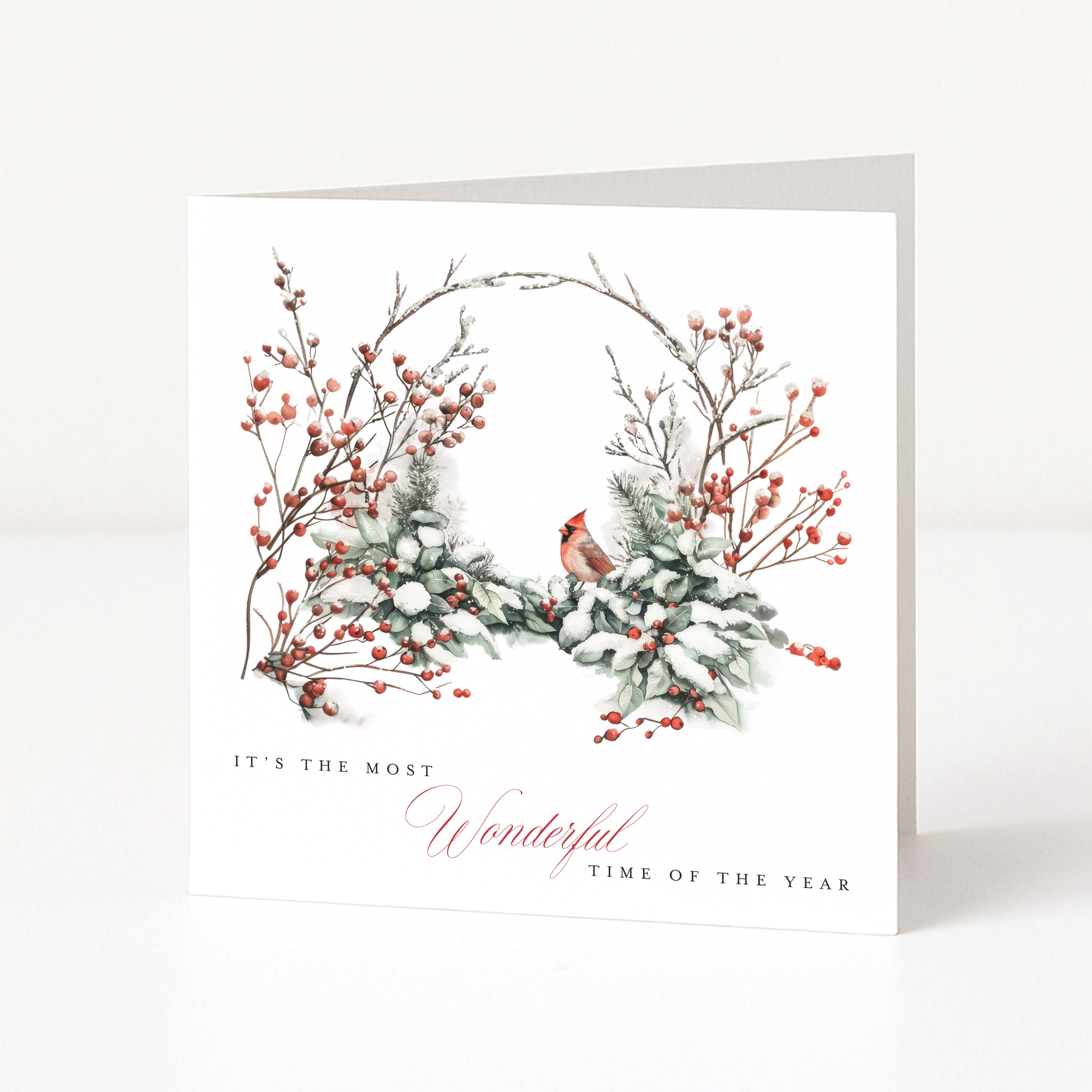 A white greeting card standing upright on a white surface, featuring a watercolour-style winter wreath with red berries, snowy greenery, a small red cardinal, and the text 'IT'S THE MOST Wonderful TIME OF THE YEAR'.