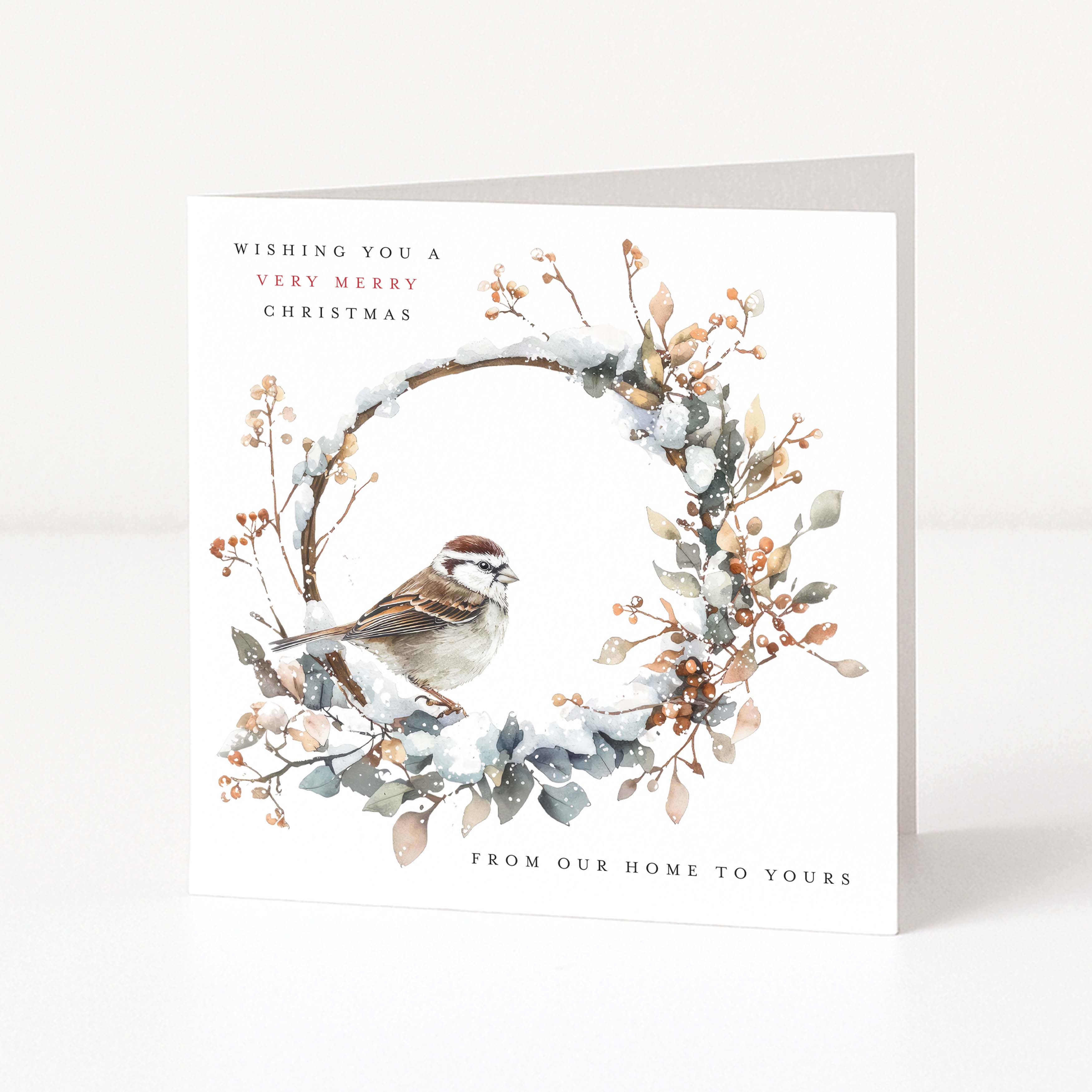 A square Christmas greeting card standing upright on a white surface. The card features a watercolour-style wreath with snow, leaves, berries, and a small bird, with the text 'Wishing you a very merry christmas' and 'From our home to yours'.