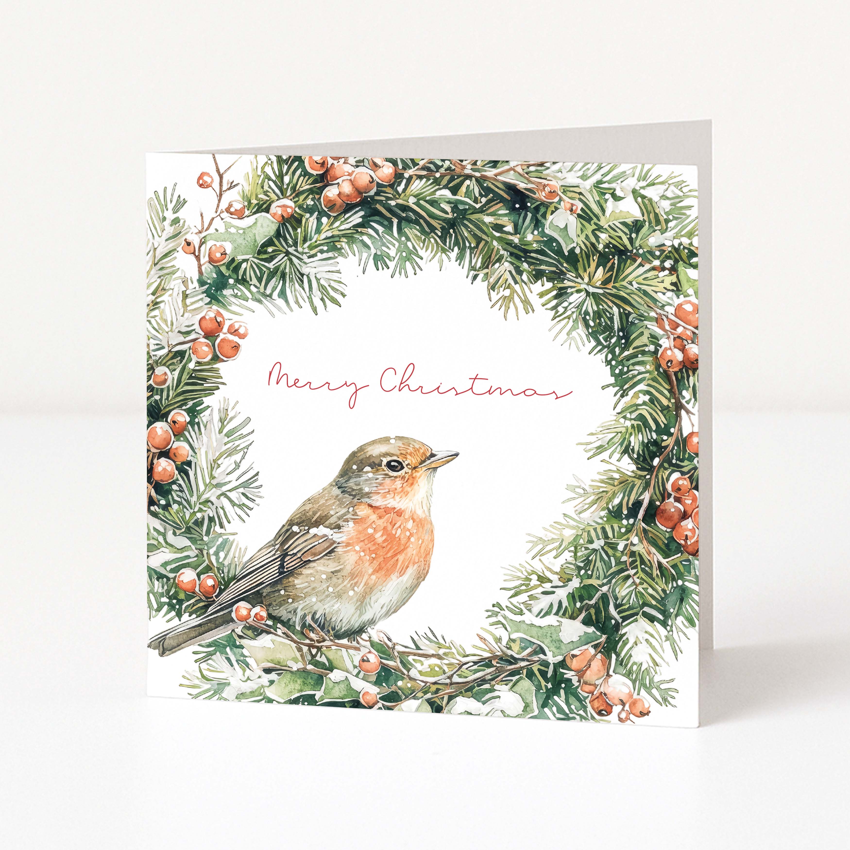 A square Christmas greeting card featuring a watercolor robin bird framed by a green pine wreath with red berries, on a white background with the words 'Merry Christmas' in pink script.
