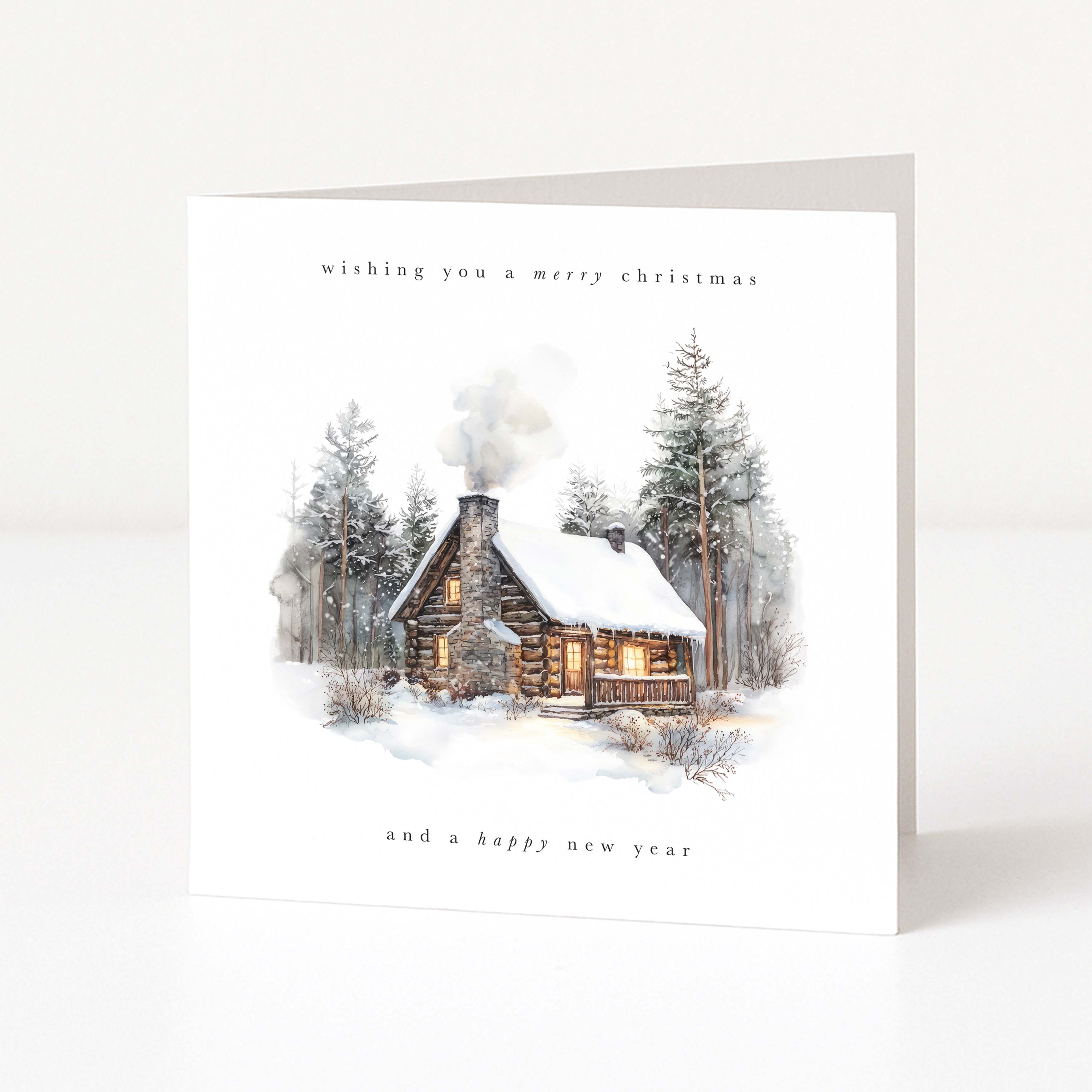 A square white Christmas greeting card standing upright on a light neutral surface. The front features an illustrated winter cabin with snow, evergreen trees, and soft smoke, with the text 'wishing you a merry christmas' at the top and 'and a happy new year' at the bottom.