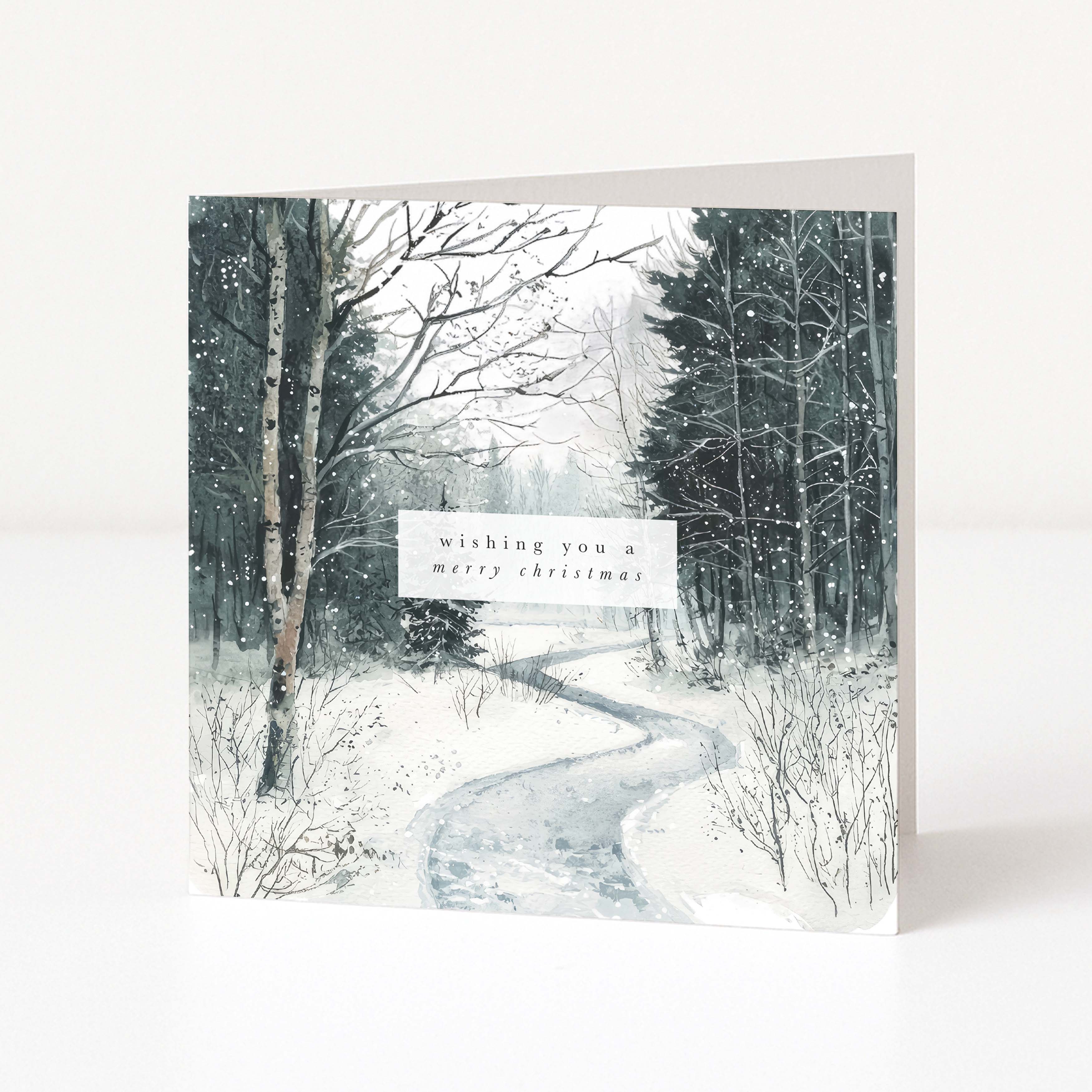 A square Christmas greeting card standing upright on a white surface, printed with a snowy forest path scene and a small white label reading 'wishing you a merry christmas'.