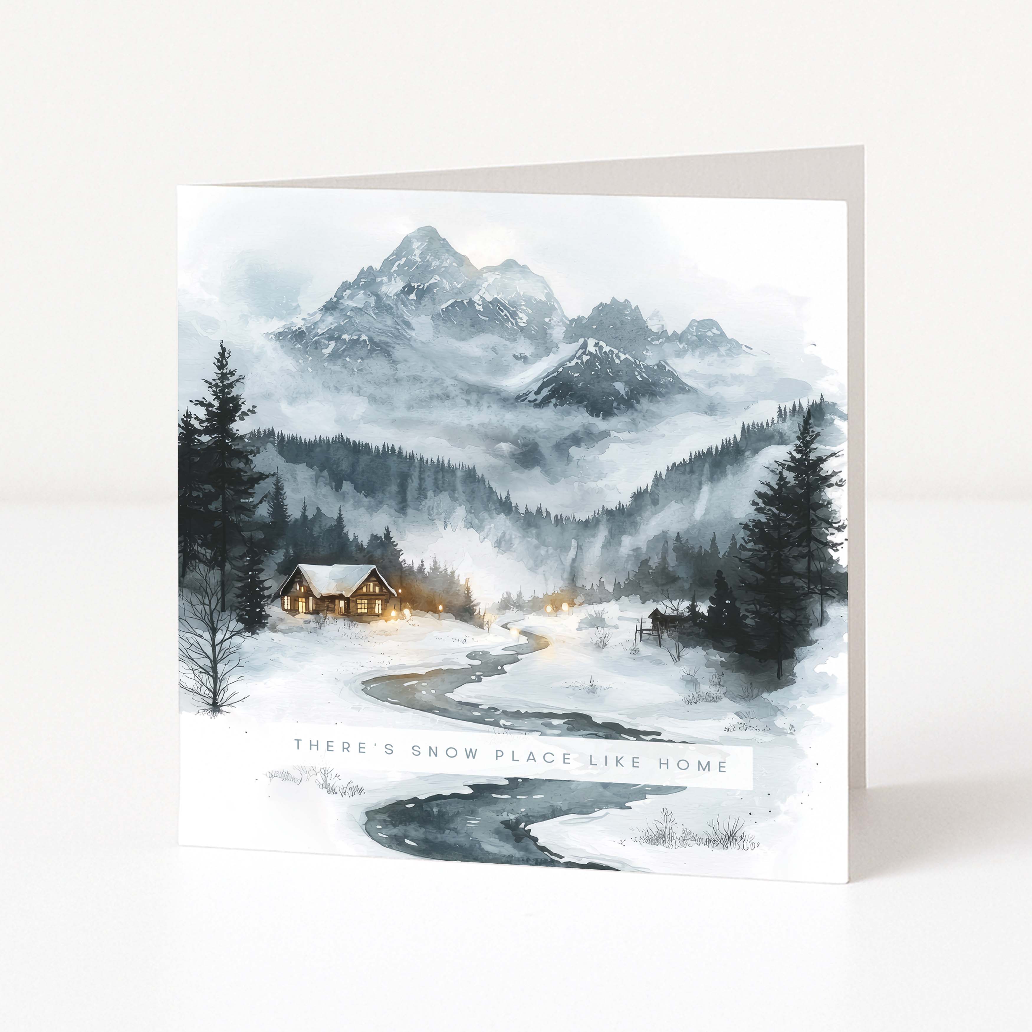 A square Christmas greeting card featuring a winter mountain landscape illustration with snow-covered peaks, dark evergreen trees, a winding road, and a small warmly lit cabin. The card is photographed standing upright against a clean white background.