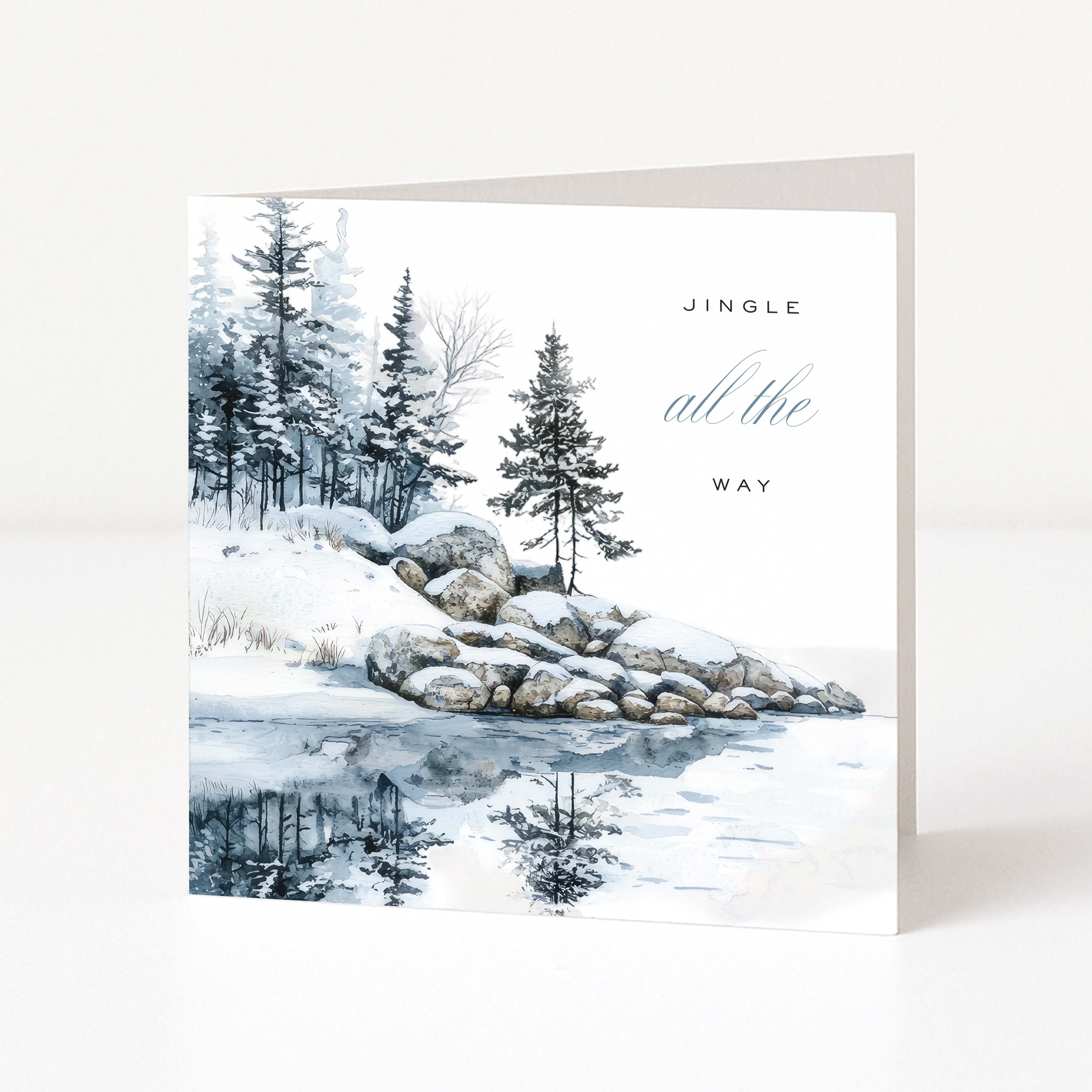 A square Christmas greeting card with a winter landscape illustration, featuring snow-covered rocks, evergreen trees, and a reflective icy surface. The card reads 'JINGLE all the WAY' in black and blue-gray text.