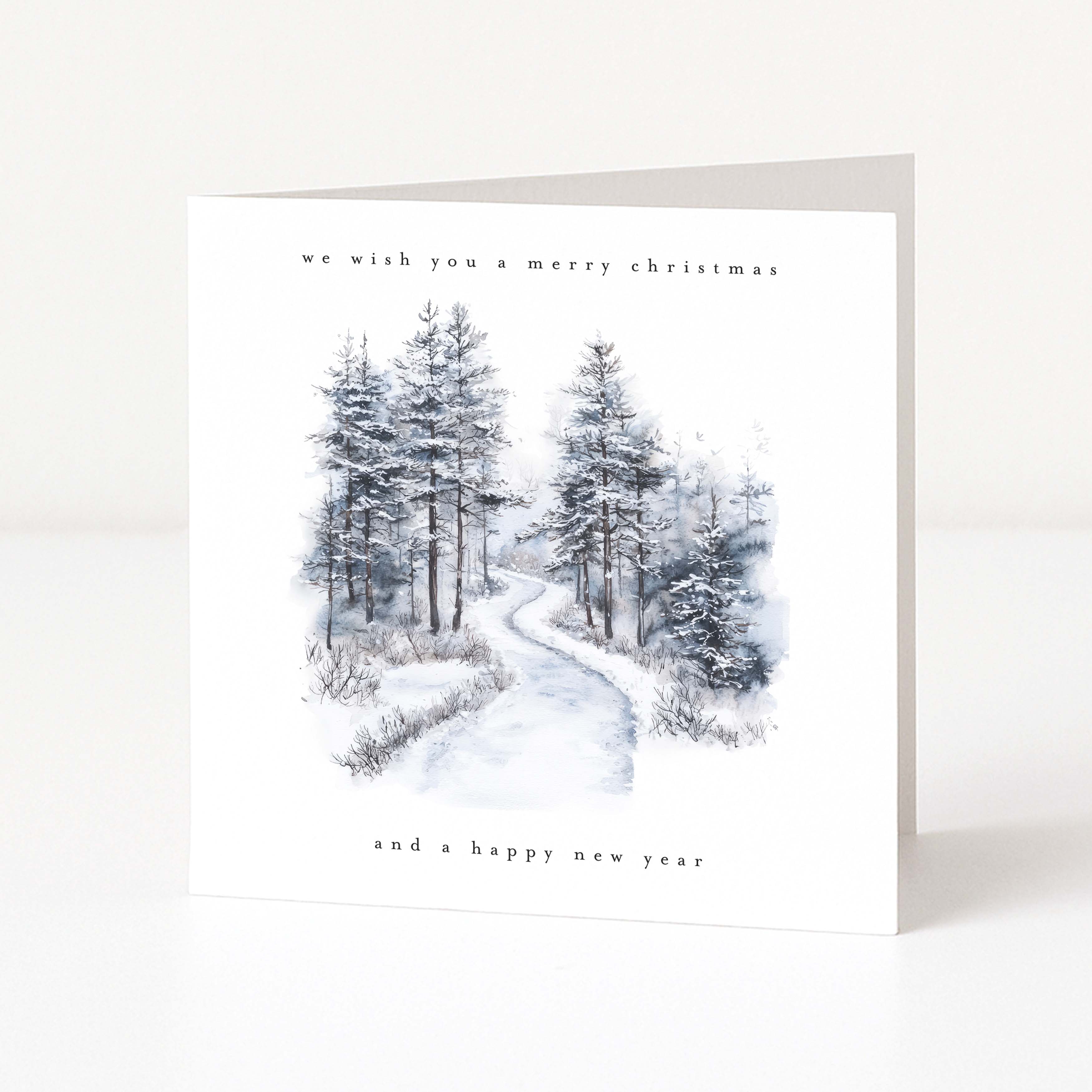 A square white Christmas greeting card standing upright on a light neutral surface. The front features a watercolor-style winter forest scene with snow-covered evergreen trees and a winding path, with the text 'we wish you a merry christmas' at the top and 'and a happy new year' at the bottom.