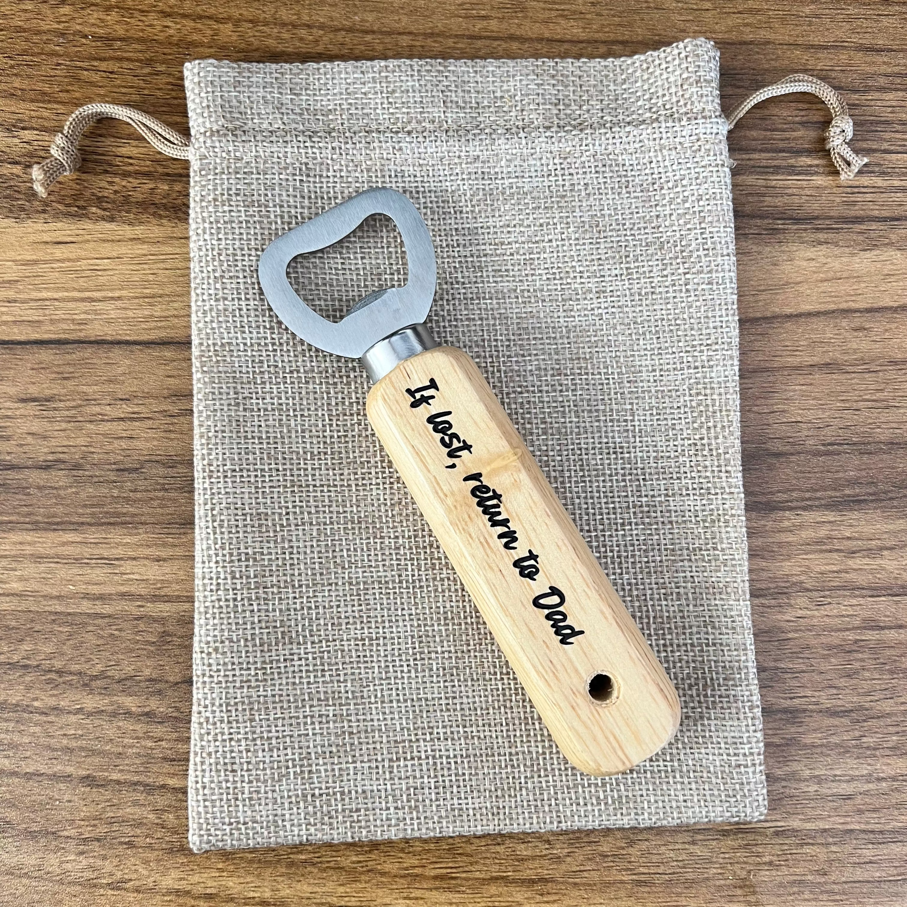 Wooden bottle opener with engraved text on a fabric pouch, placed on a wooden surface.