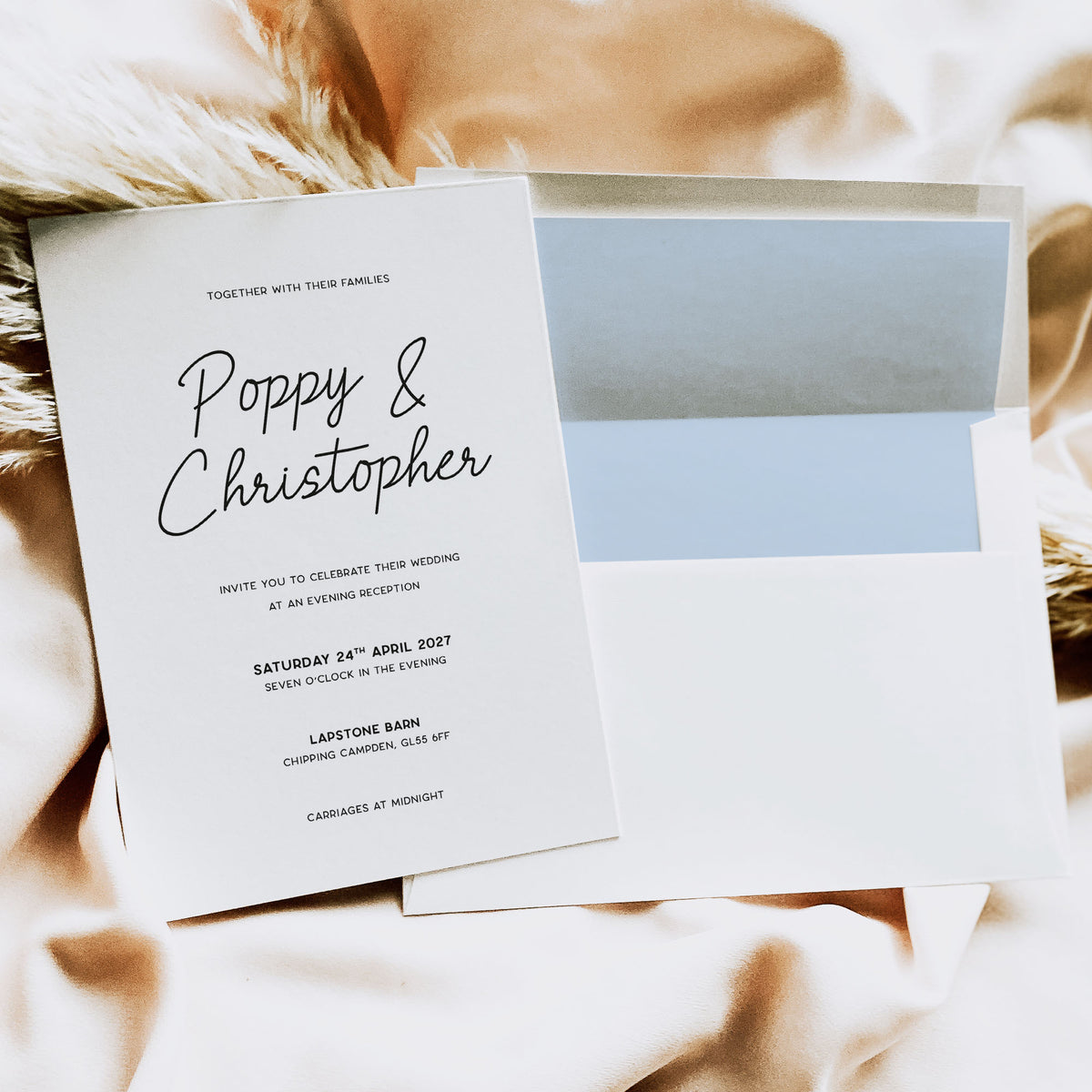A wedding invitation card and matching envelope laid on soft beige fabric. The white card has black script and serif text reading 'Poppy & Christopher' with wedding details below, and the envelope has a pale blue center panel.