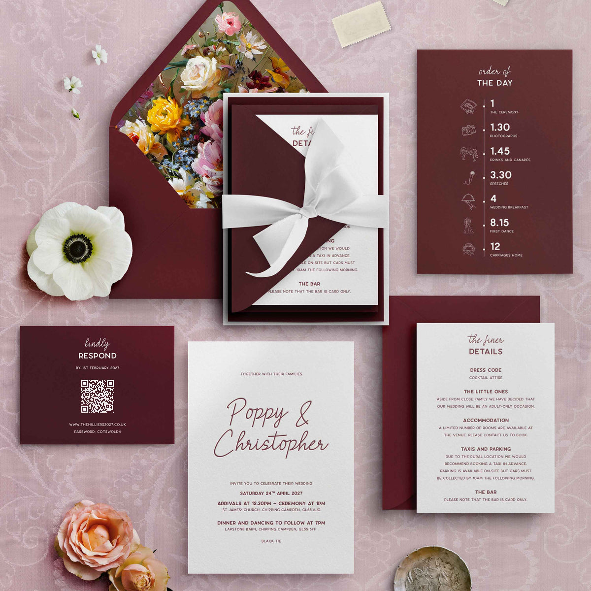 A styled flat lay of a wedding invitation suite on a dusty pink patterned background, including a burgundy letterpress and white invitation card tied with a white ribbon, a floral envelope, a details card, a RSVP card, and decorative flowers.