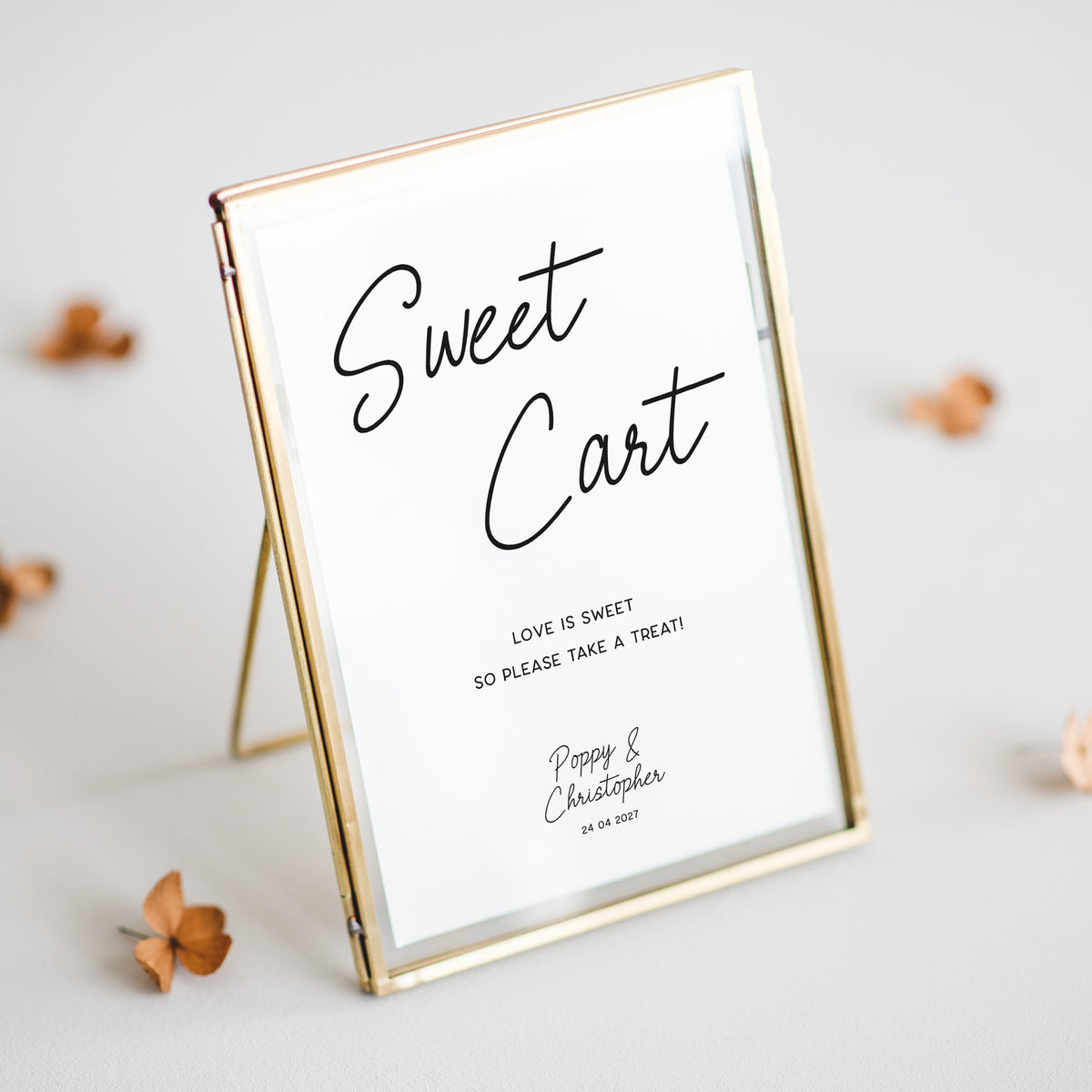 A rectangular picture frame with a gold-toned border and A-frame stand, holding a white sign that reads 'Sweet Cart' with smaller wedding text, photographed on a light neutral surface with a few dried flowers around it.