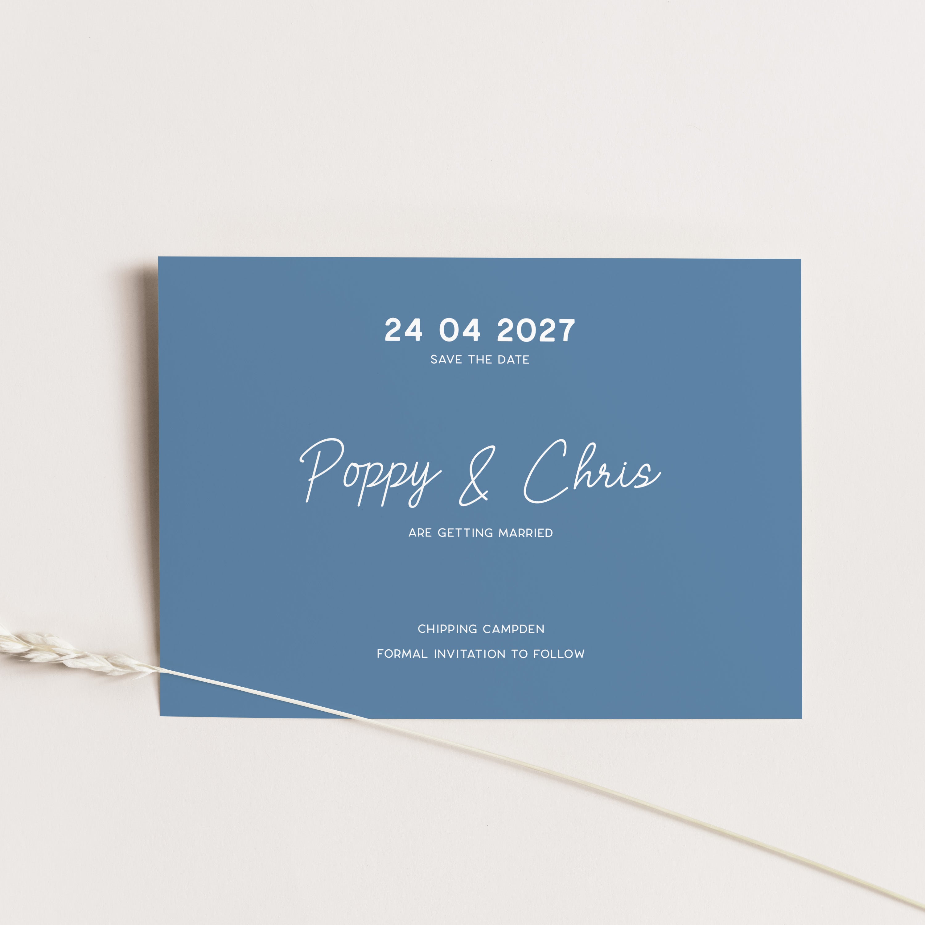 A rectangular blue wedding save-the-date card with white text, photographed on a light neutral background with a thin decorative plant stem crossing the lower left corner.