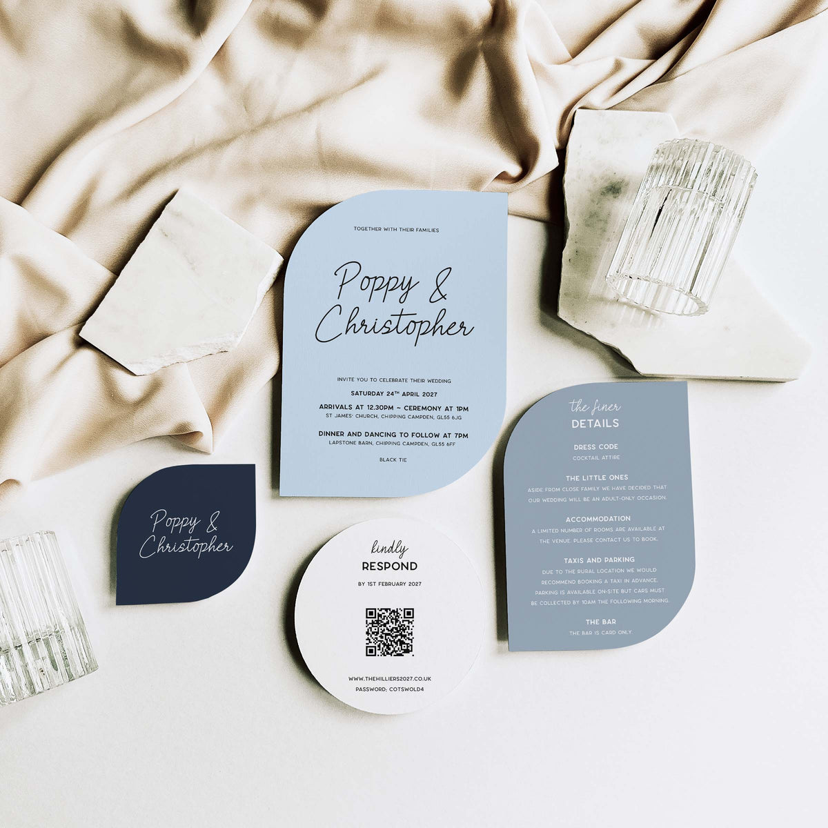 A styled flat-lay of wedding stationery on a white surface with draped beige fabric. The set includes a light blue invitation card, a dark navy badge, a white round card with a QR code, and a muted blue details card, arranged with glassware and paper clips.