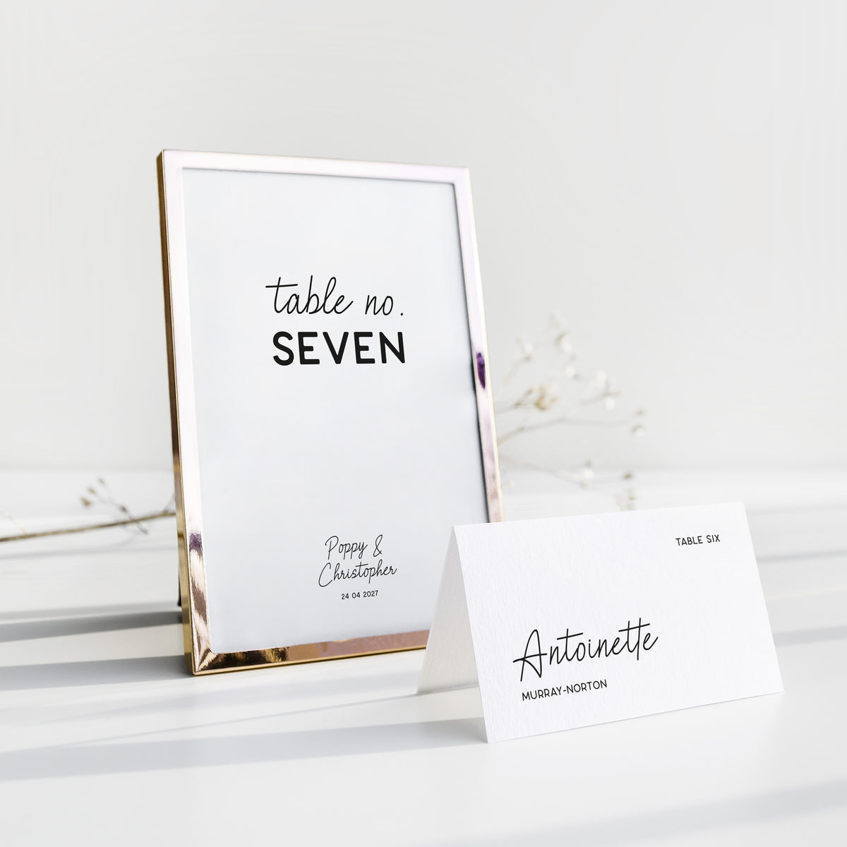 A wedding table number in a gold frame next to a folded place card on a white background