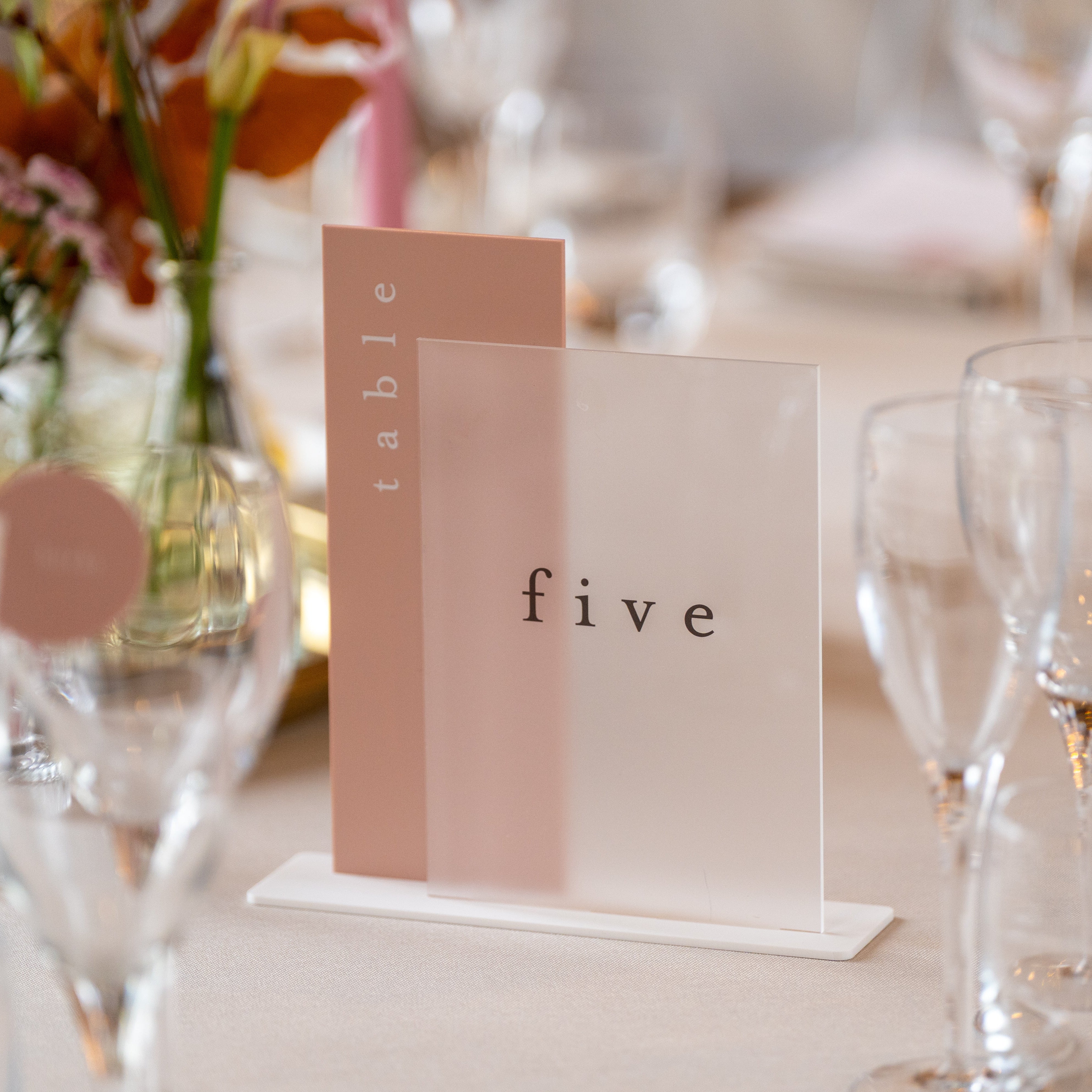 Table number 'five' on a stand with floral decorations and glasses in the background