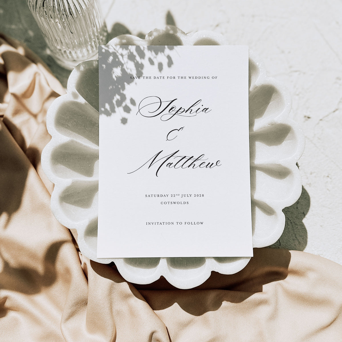 A white save the date card on both a marble background and a white seashell style plate alongside neutral fabric
