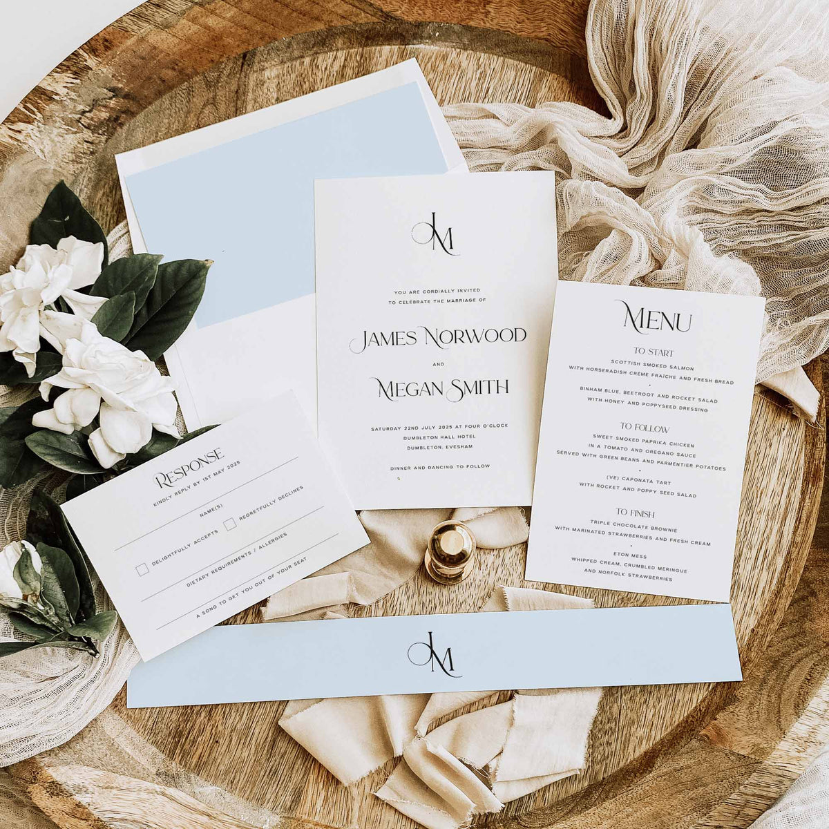 A wedding invitation flat lay with invitation, menu card, response card, pale blue monogram belly band and a white straight flap envelope with a pale blue envelope liner, sat amongst neutral fabric and foliage.