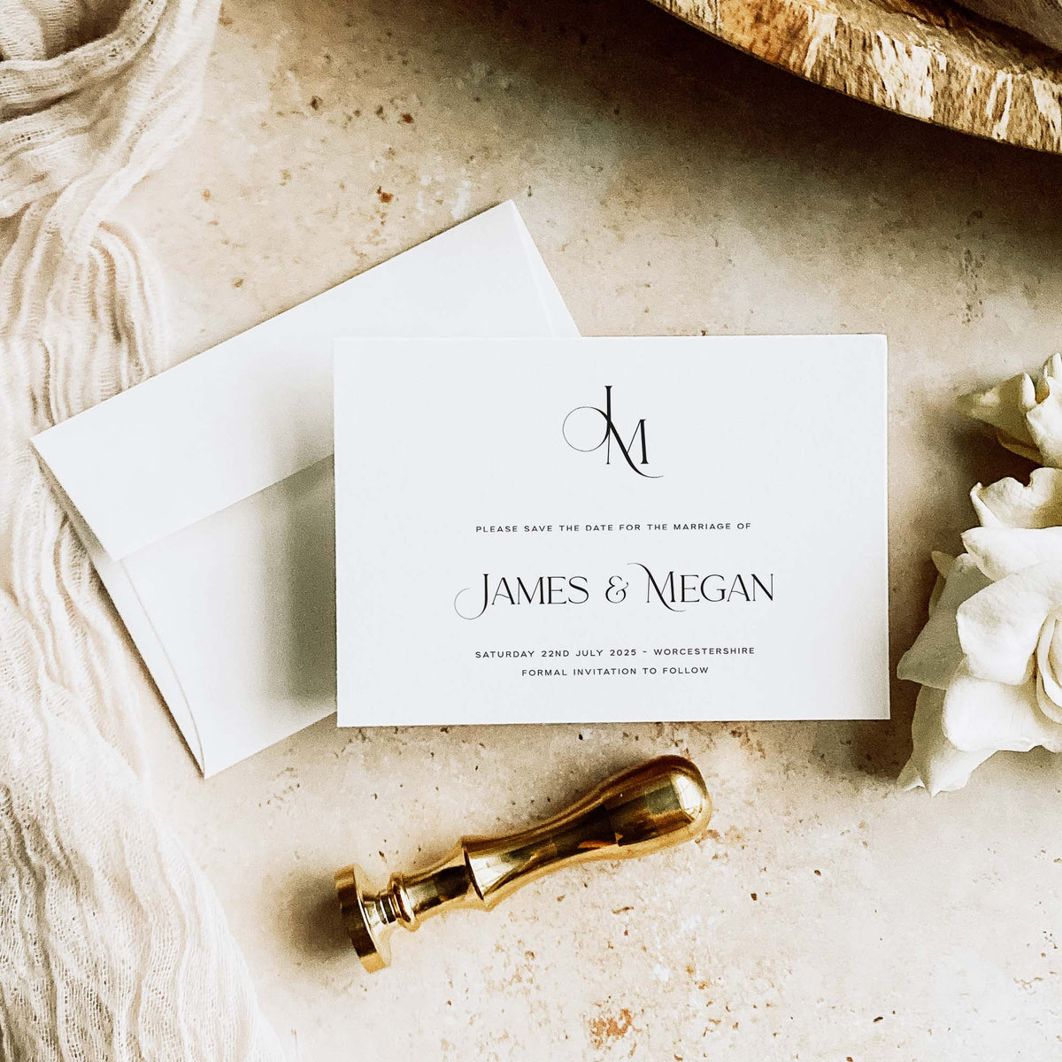 A white save the date card for a wedding on a neautral marble background, shown with the back of a white envelope, wax stamp and white fabric.