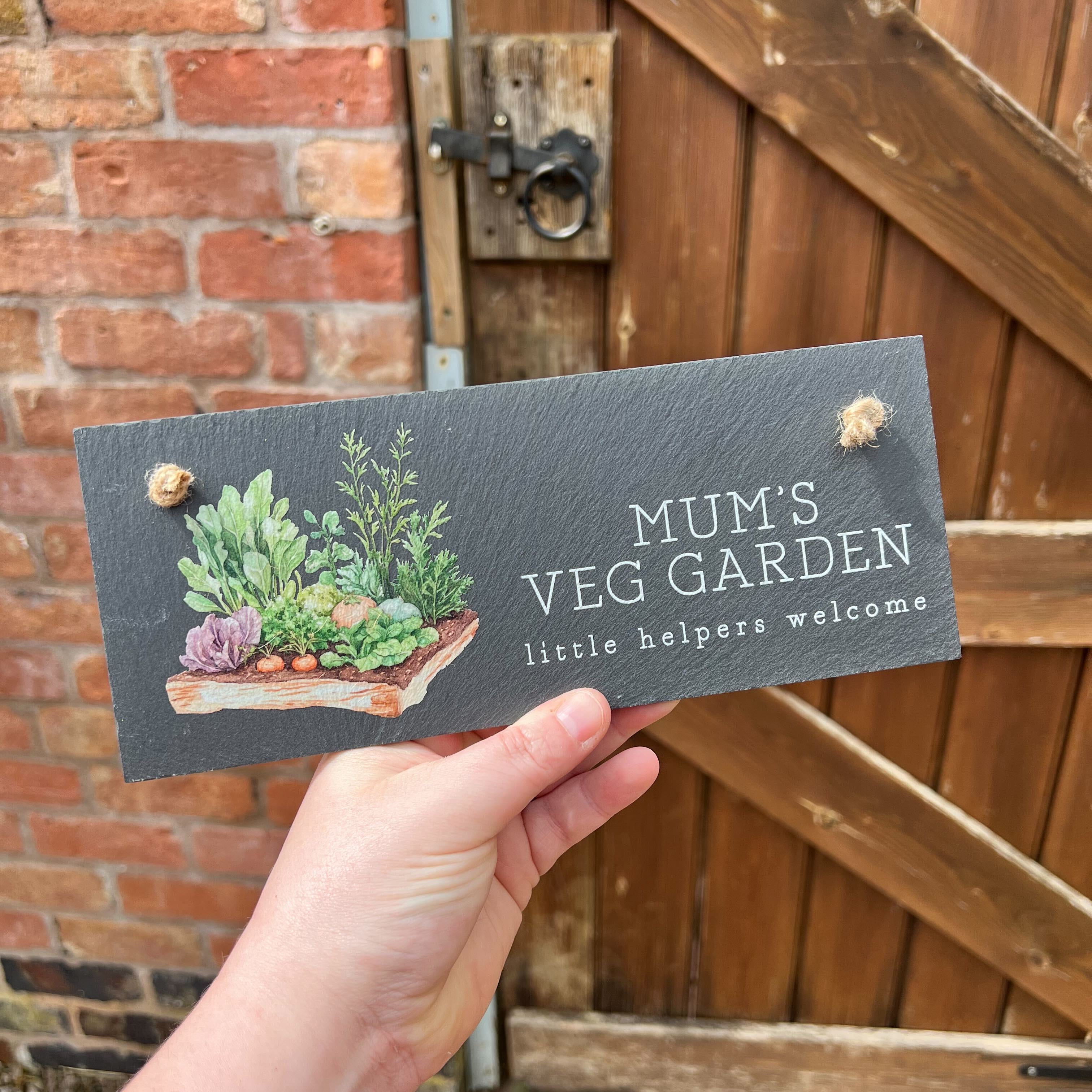 Hand holding a sign with 'Mum's Veg Garden' and plant illustrations in front of a wooden gate.