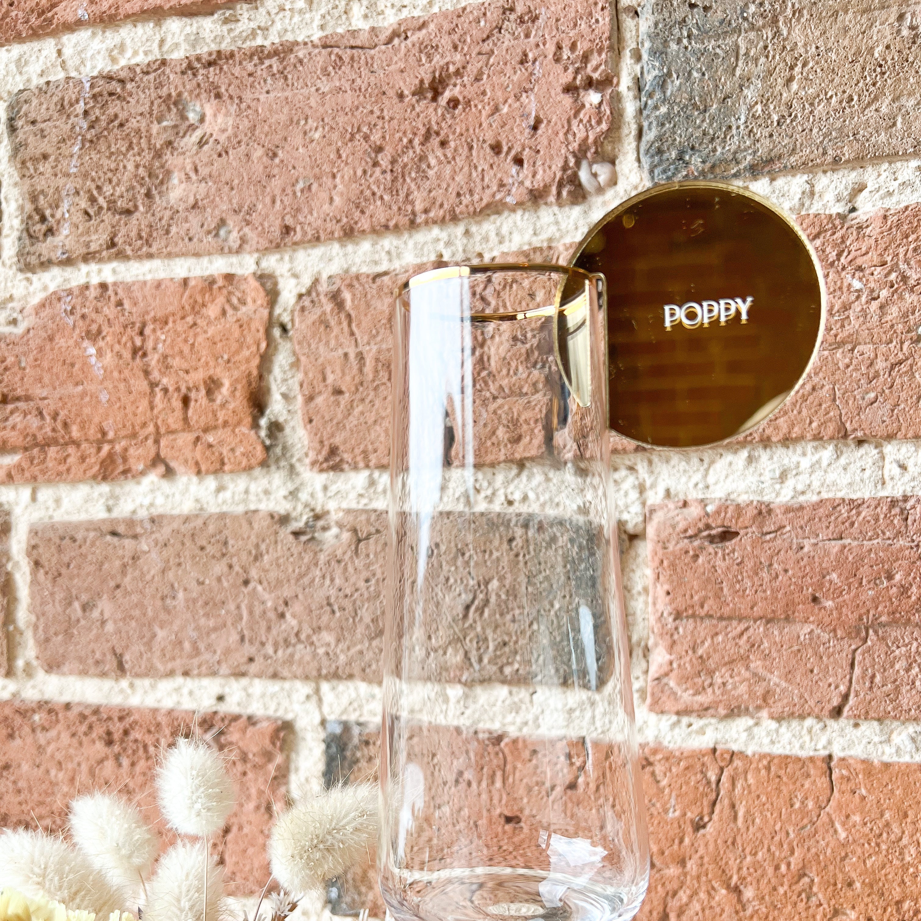 Clear glass with gold drink token on rim labeled 'Poppy' against a brick wall.
