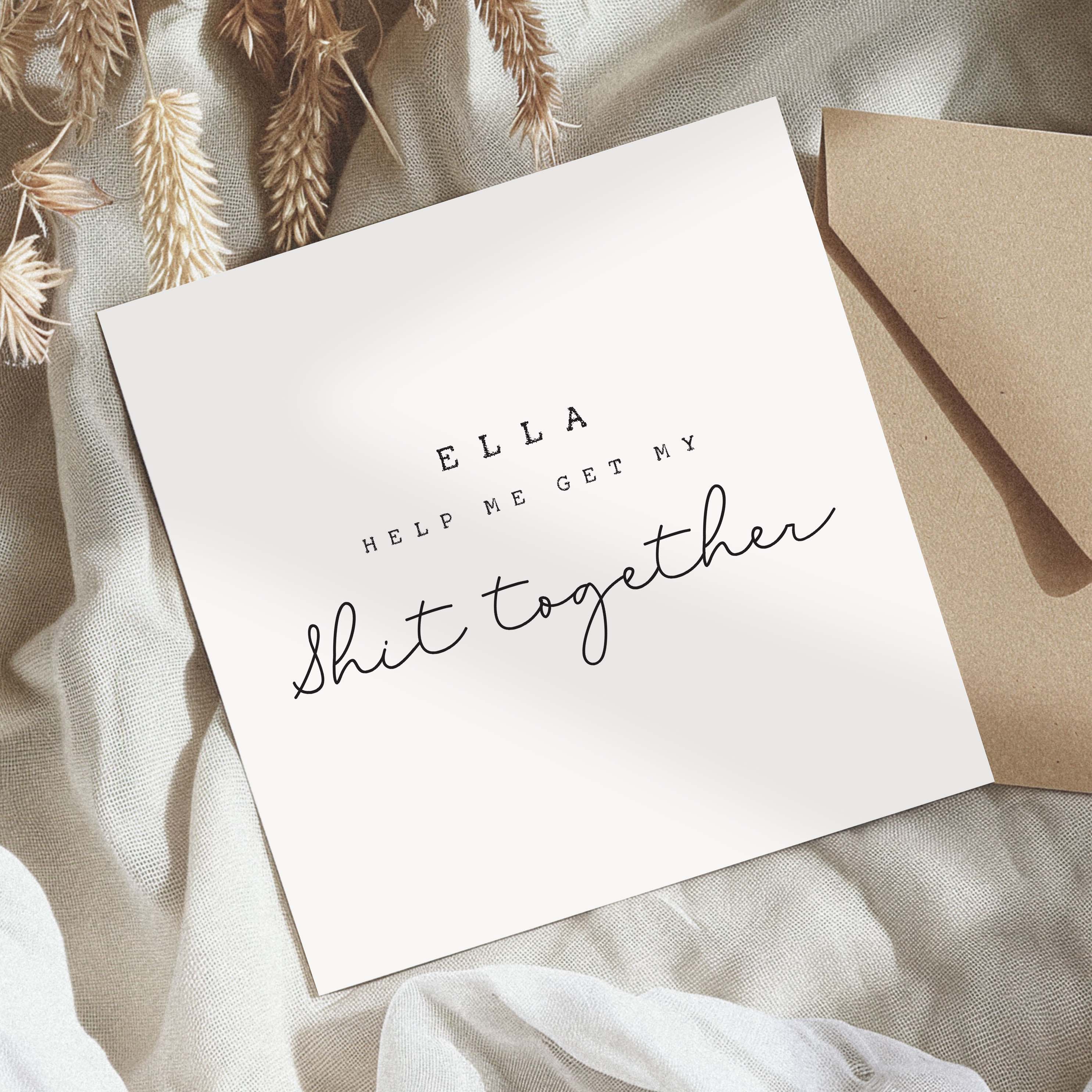 White card with text 'ELLA HELP ME GET MY SHIT TOGETHER' on a textured surface with an envelope.