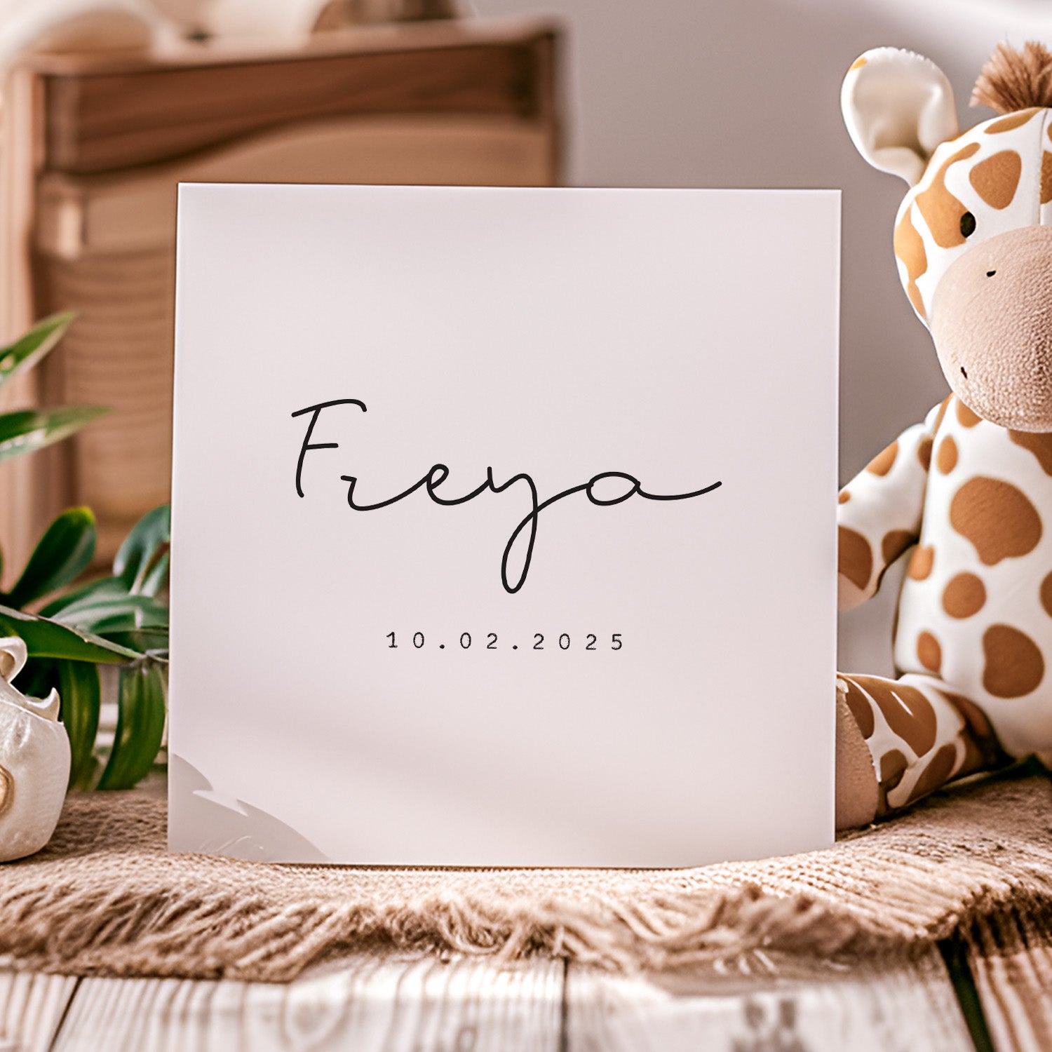White card with 'Freya' and date on a wooden surface with a giraffe plush toy and plant in the background