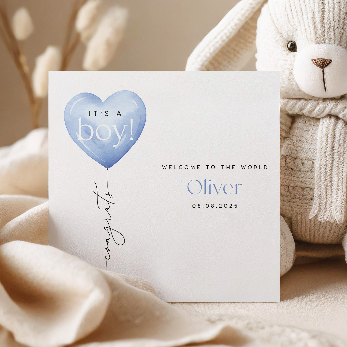 Greeting card for a baby boy with a blue heart balloon, surrounded by soft textures and a teddy bear.