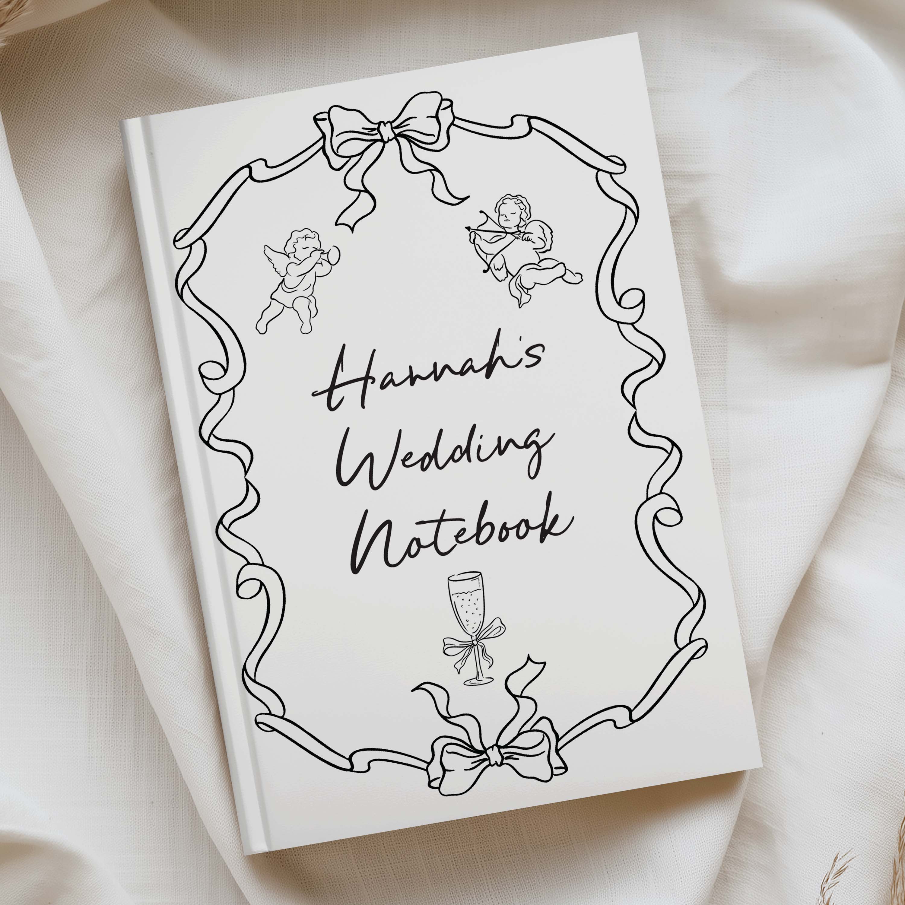 A hardcover wedding notebook titled 'Hannah's Wedding Notebook' in black script, decorated with line-art illustrations of cherubs, ribbons, and a champagne glass. The notebook is photographed on draped cream fabric.