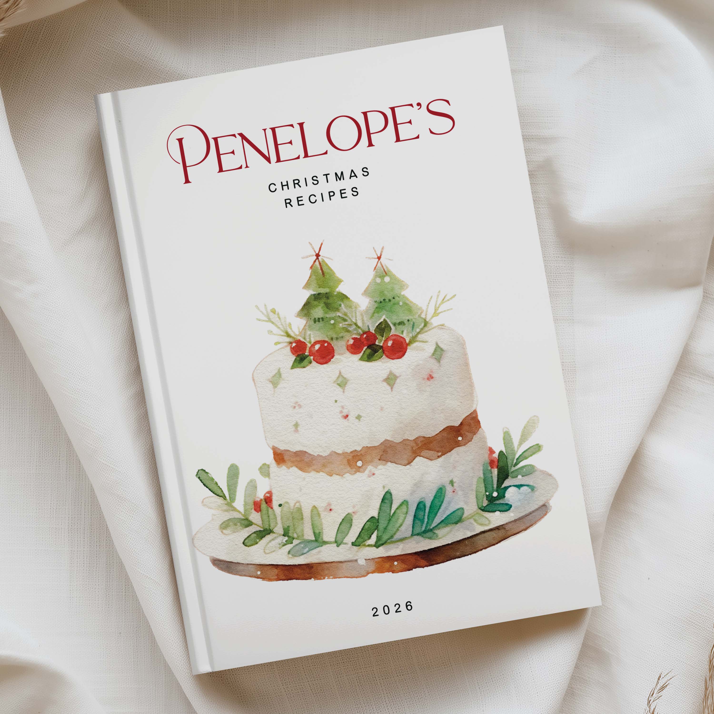 A hardcover cookbook titled 'PENELOPE'S CHRISTMAS RECIPES 2026' resting on a draped white fabric background. The cover features a watercolor illustration of a decorated Christmas cake with green holly leaves and red berries.
