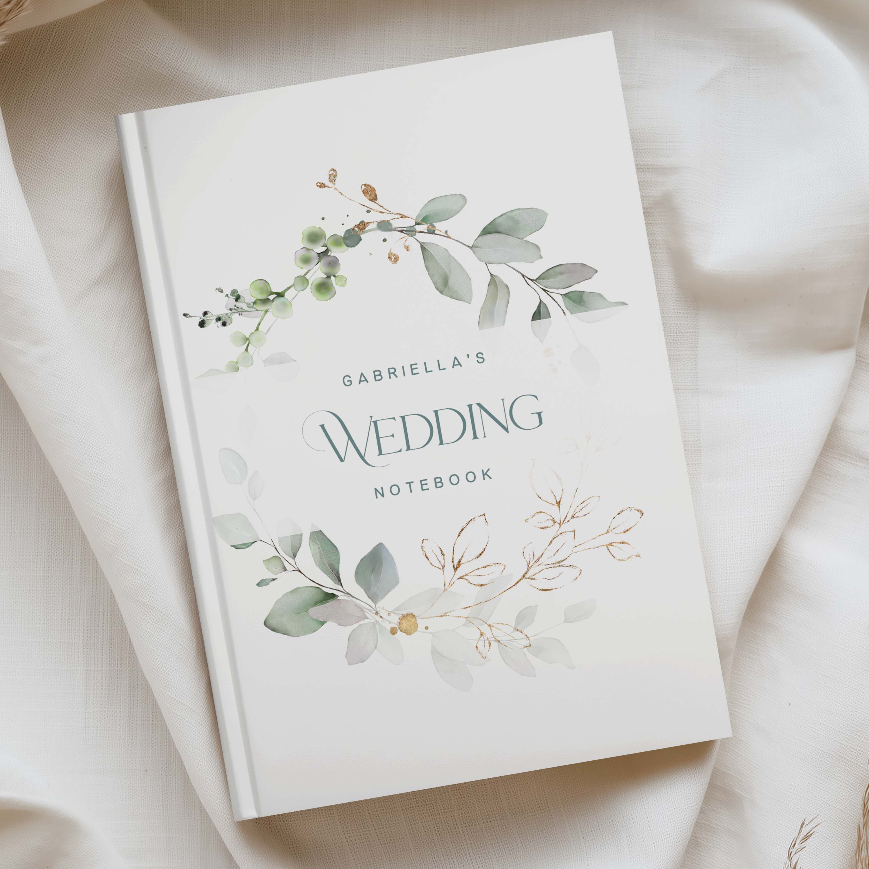 A personalized wedding notebook with a white cover, featuring watercolor green and gold botanical leaf designs and the text 'GABRIELLA'S WEDDING NOTEBOOK' in a clean serif typeface. The notebook is photographed from an overhead angle resting on draped cream-colored fabric.