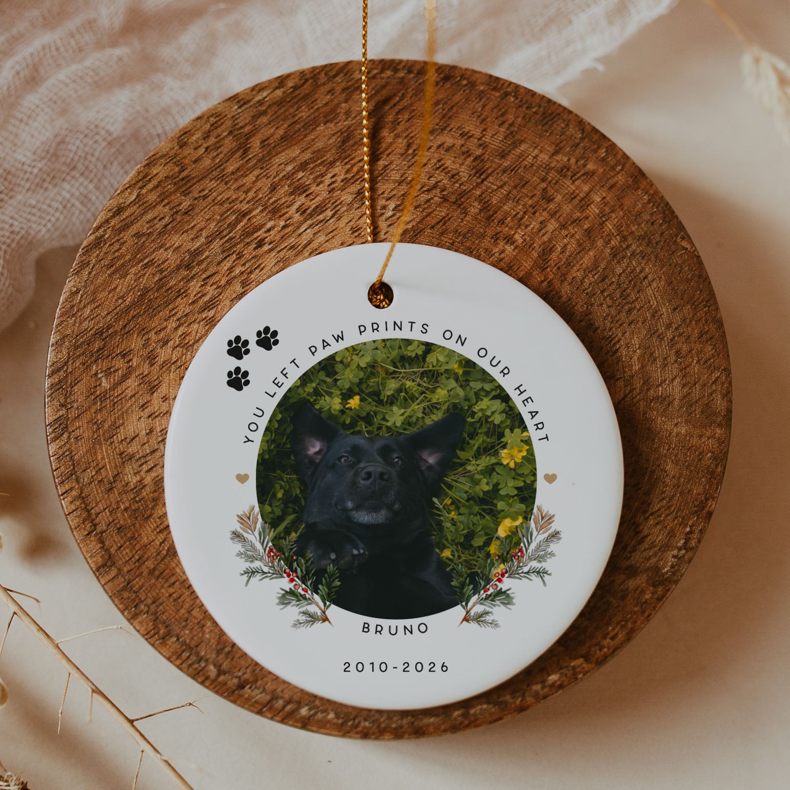 A round white memorial ornament with a hanging loop, featuring a custom photo of a black dog surrounded by greenery and floral accents, with the text 'YOU LEFT PAW PRINTS ON OUR HEART' and the name 'BRUNO' below. The ornament is displayed on a round wooden tray against a soft beige fabric background.