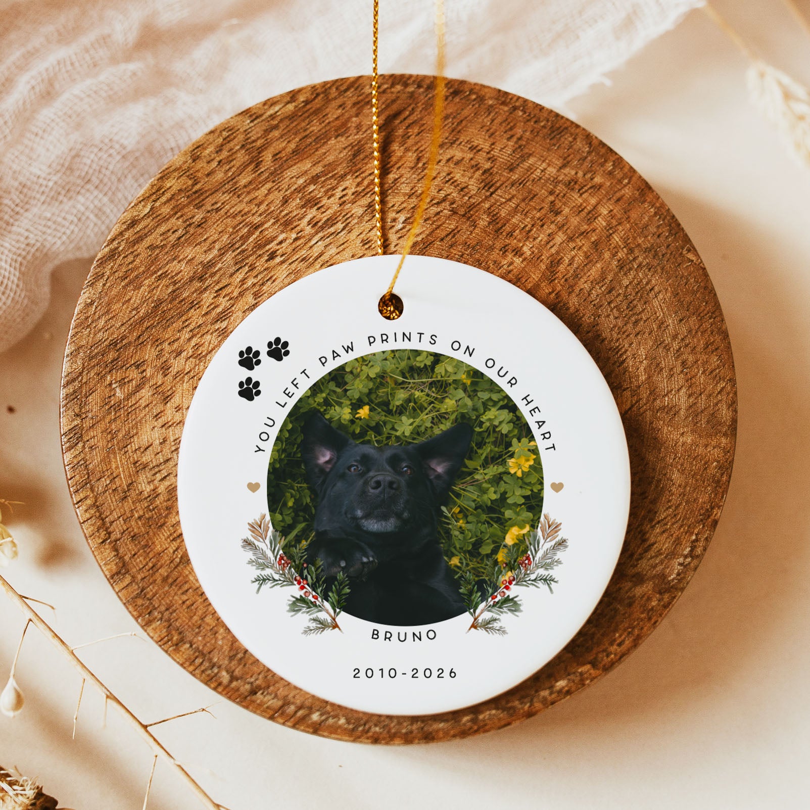 A round white personalized pet memorial ornament hanging from a gold string, displayed on a rustic wooden coaster. The ornament features a custom photo of a black dog surrounded by green foliage and yellow flowers, with the text 'YOU LEFT PAW PRINTS ON OUR HEART', the name 'BRUNO', and the years '2010-2026'.