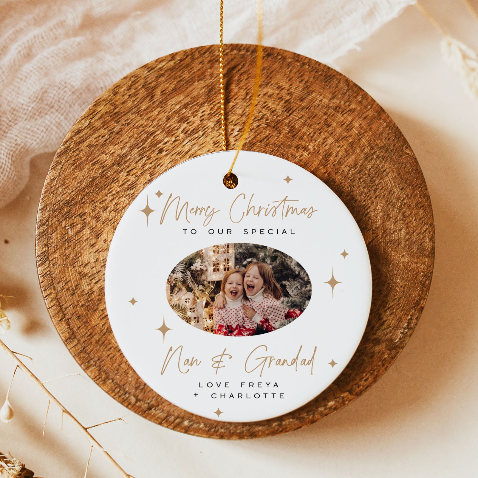 A round white personalized Christmas ornament with gold script text and star accents, displayed on a round wooden board. The ornament features a custom photo of two children and the text 'Merry Christmas to our special Nan & Grandad' and 'Love Freya + Charlotte'.